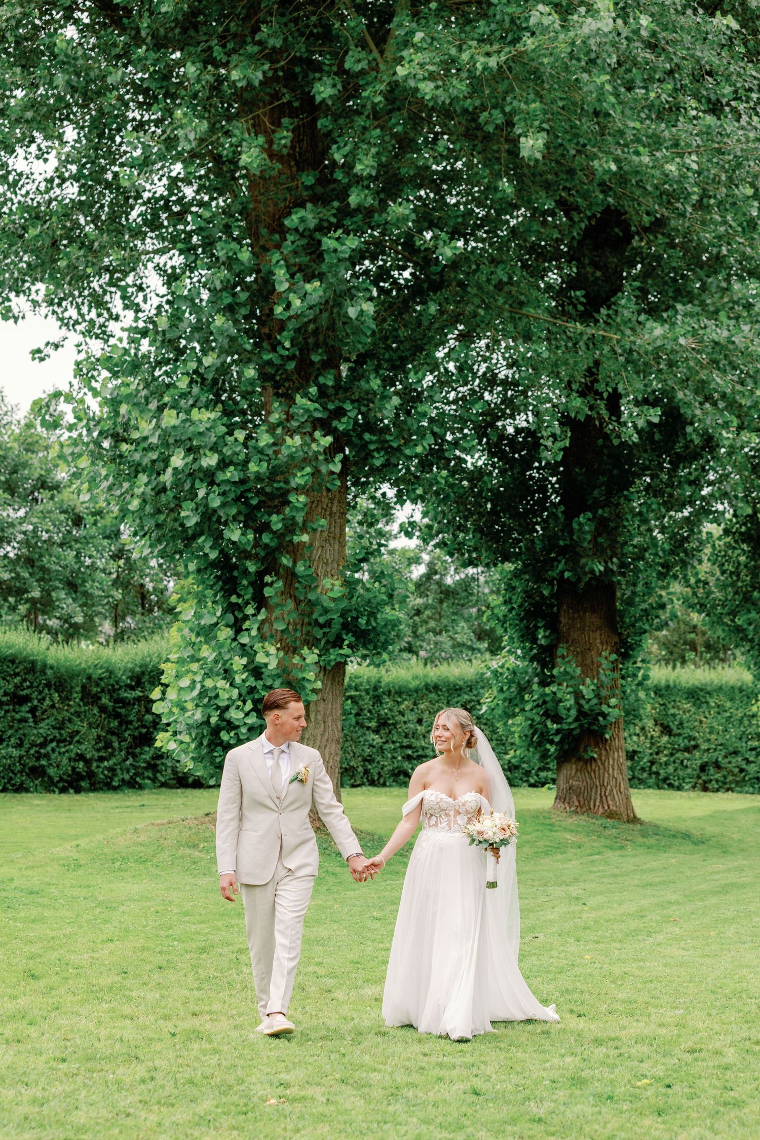 A couple in white attire walks together through a lush green garden setting on their wedding day.