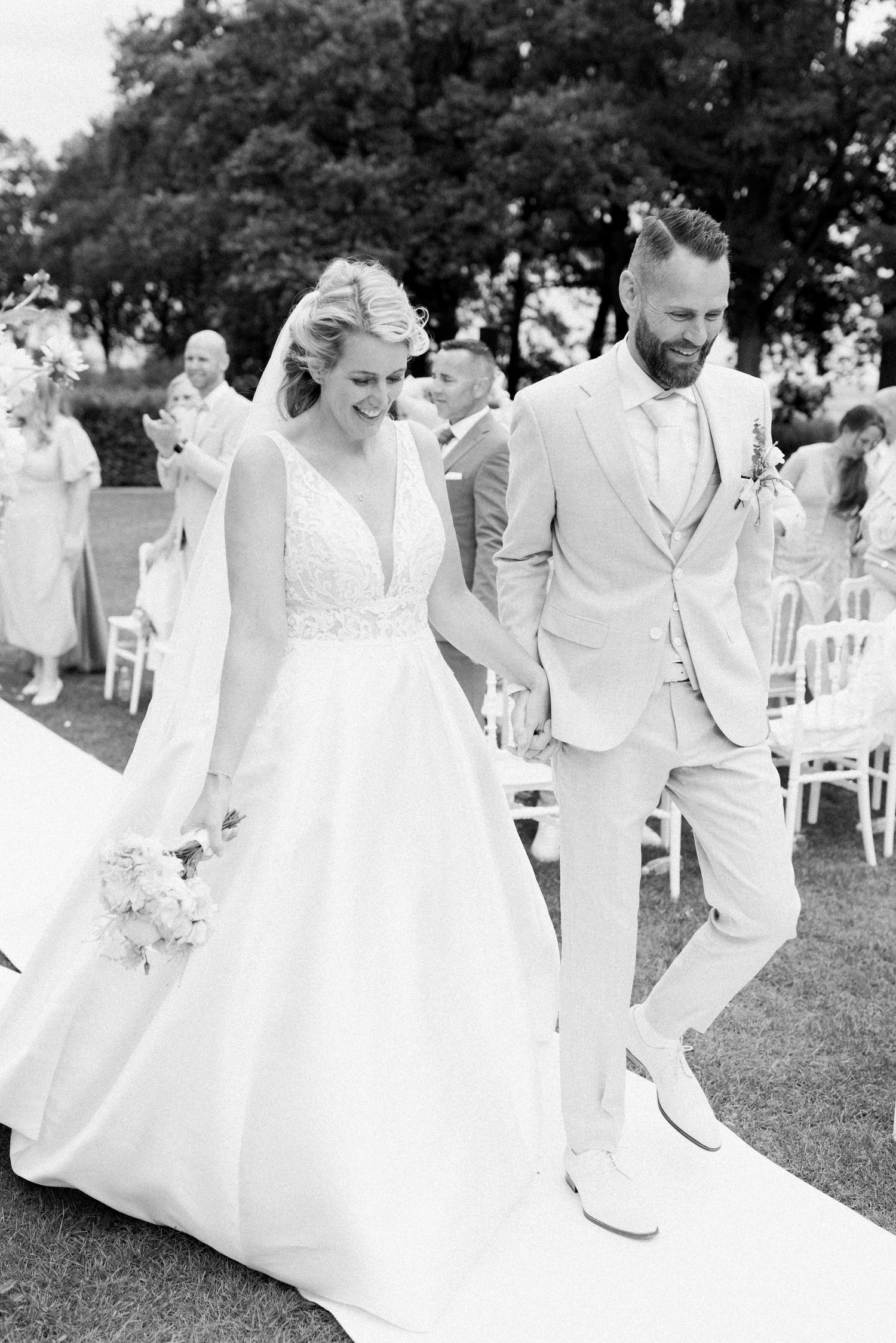 A joyful wedding couple and their bridal party all dressed in white dance and celebrate outdoors on a grassy lawn.