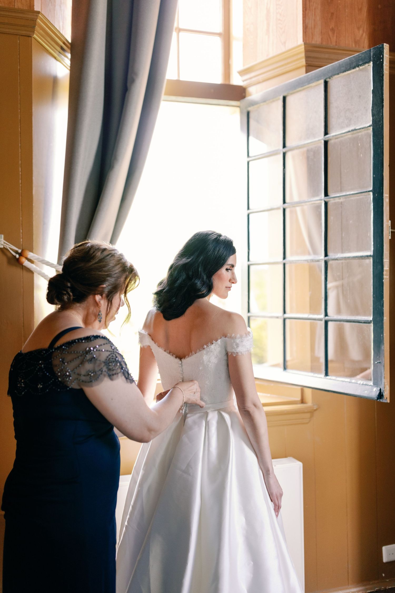 People helping with a white wedding dress by a window in a warm-lit room with yellow walls.