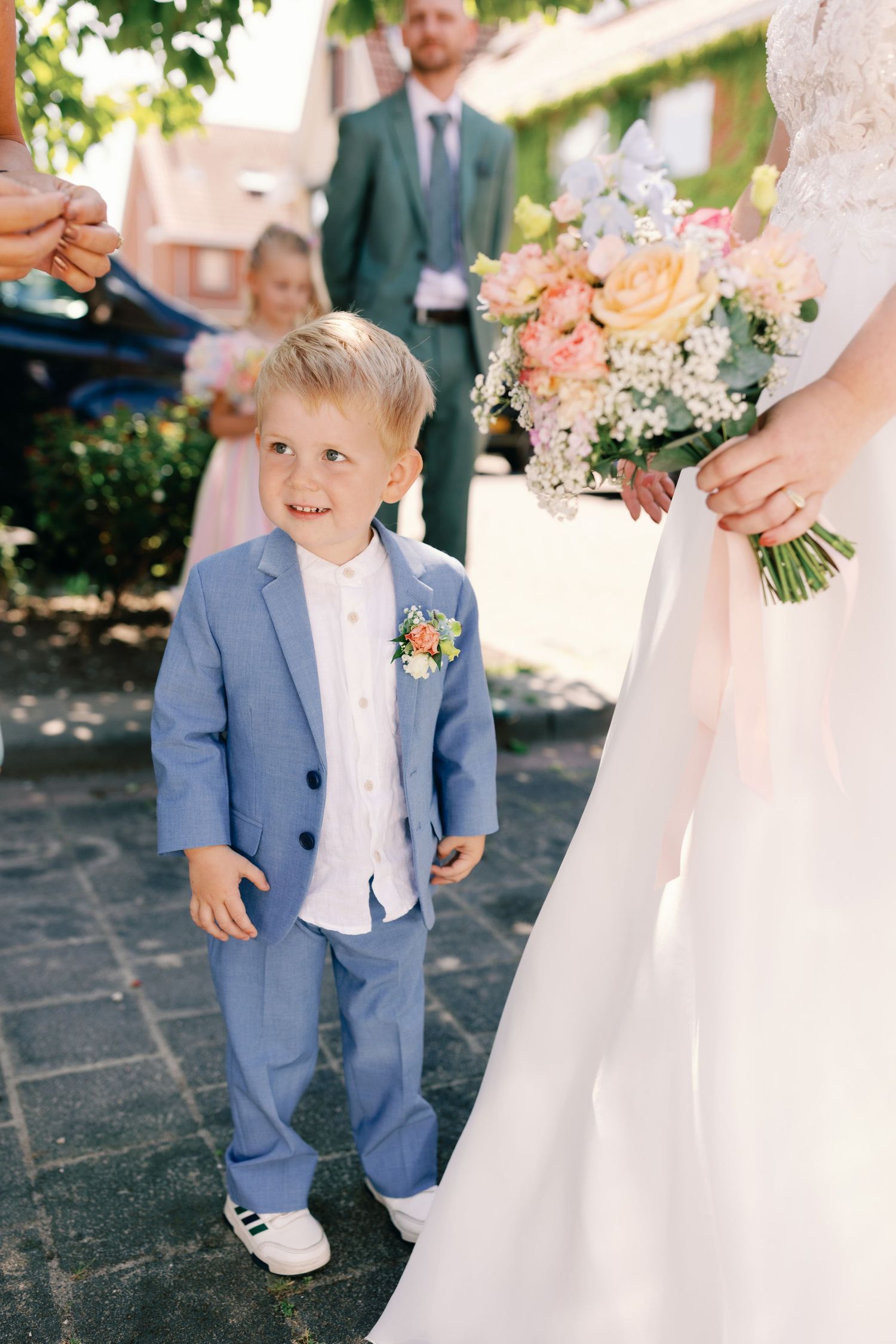 A young attendant in a light blue suit smiles while standing next to a white wedding dress at an outdoor ceremony.