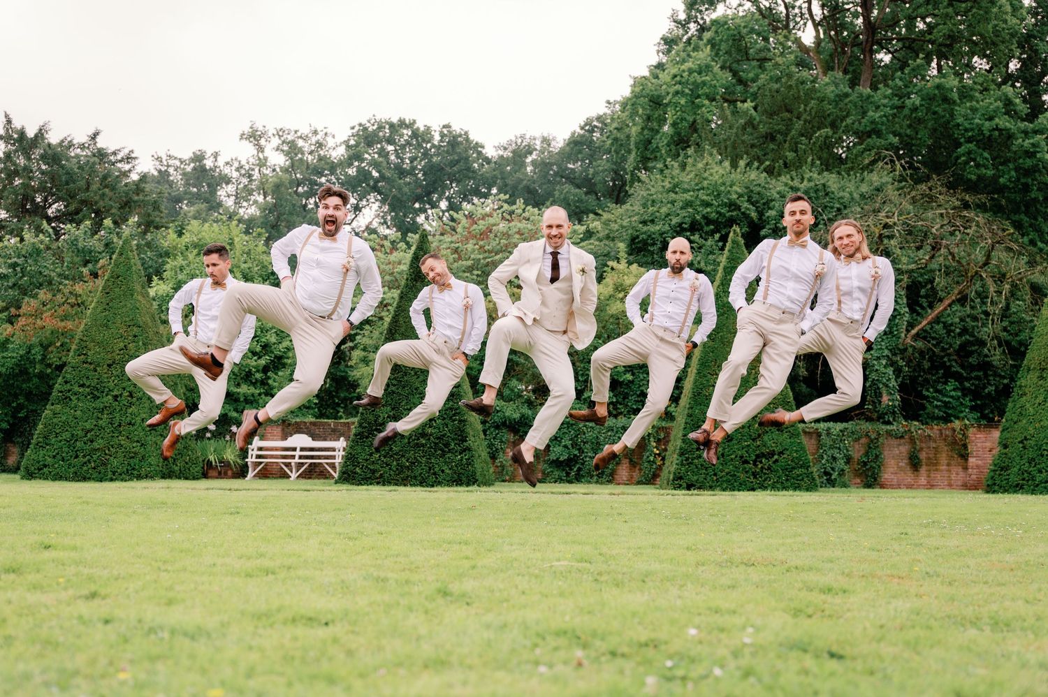 A group in matching light colored suits leaping joyfully in unison across a green lawn during an outdoor celebration.