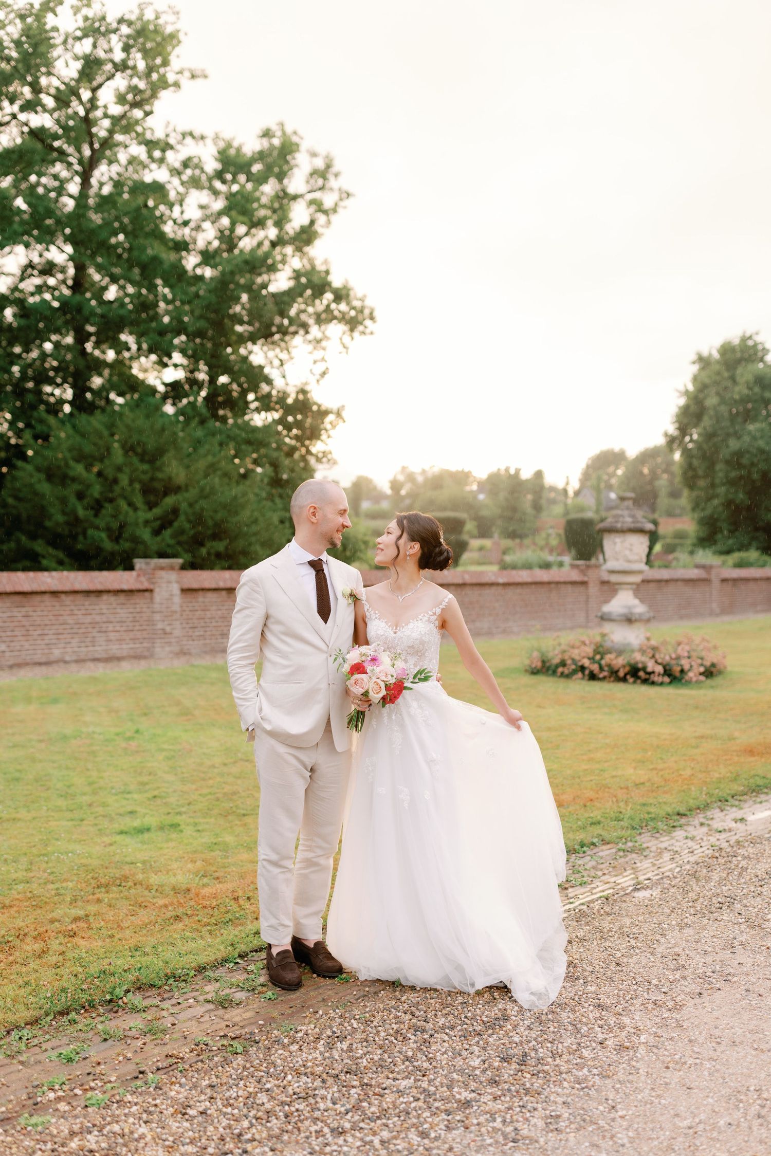 A couple in wedding attire walks along a gravel path at sunset in a garden setting.