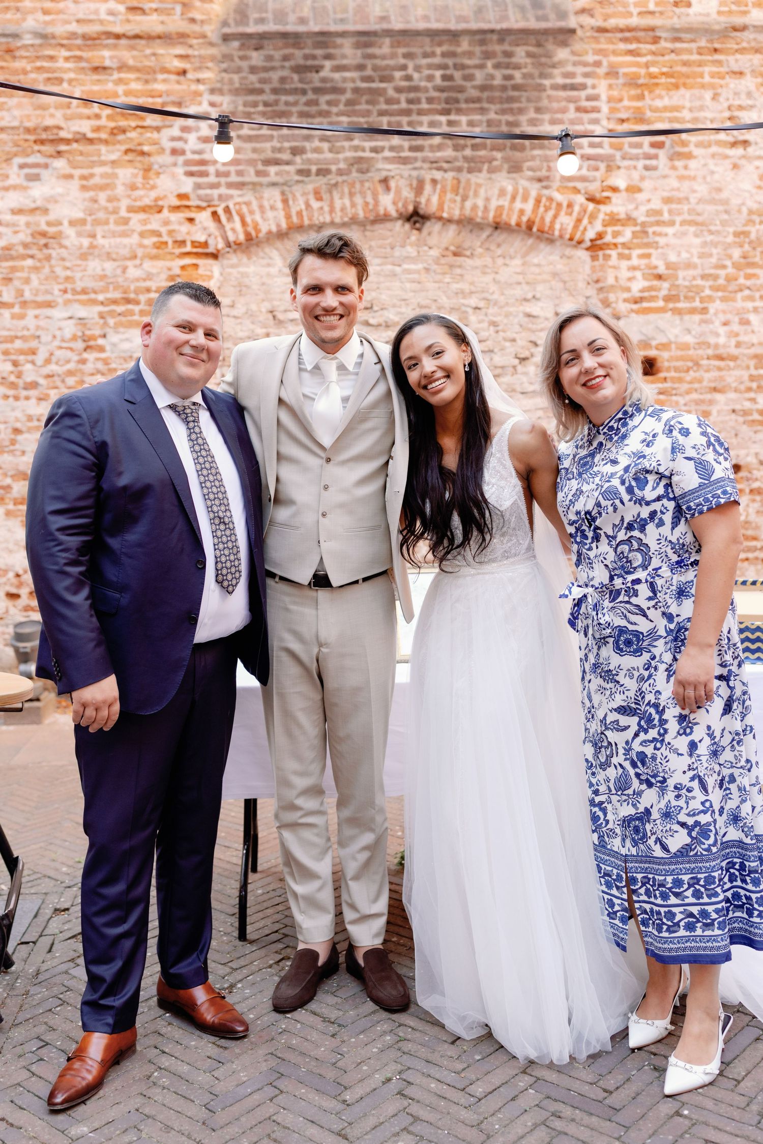 Wedding party poses against rustic brick wall in formal attire with blue and white floral dress details.
