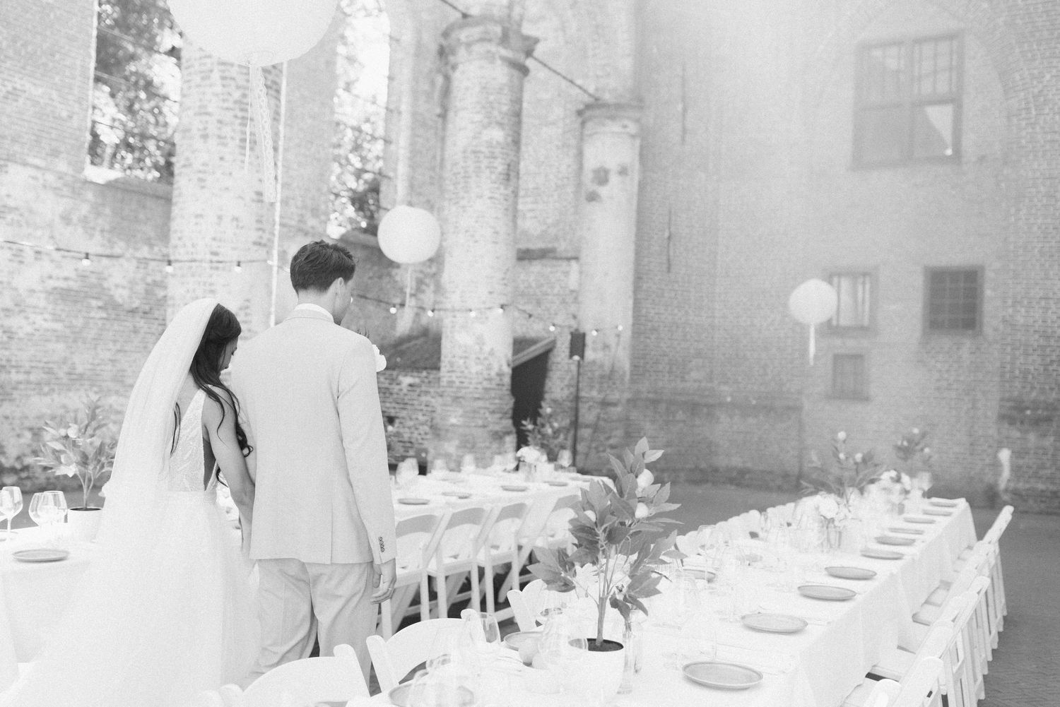 Black and white photo of an elegant outdoor wedding reception setup with long tables and white decor against stone pillars.