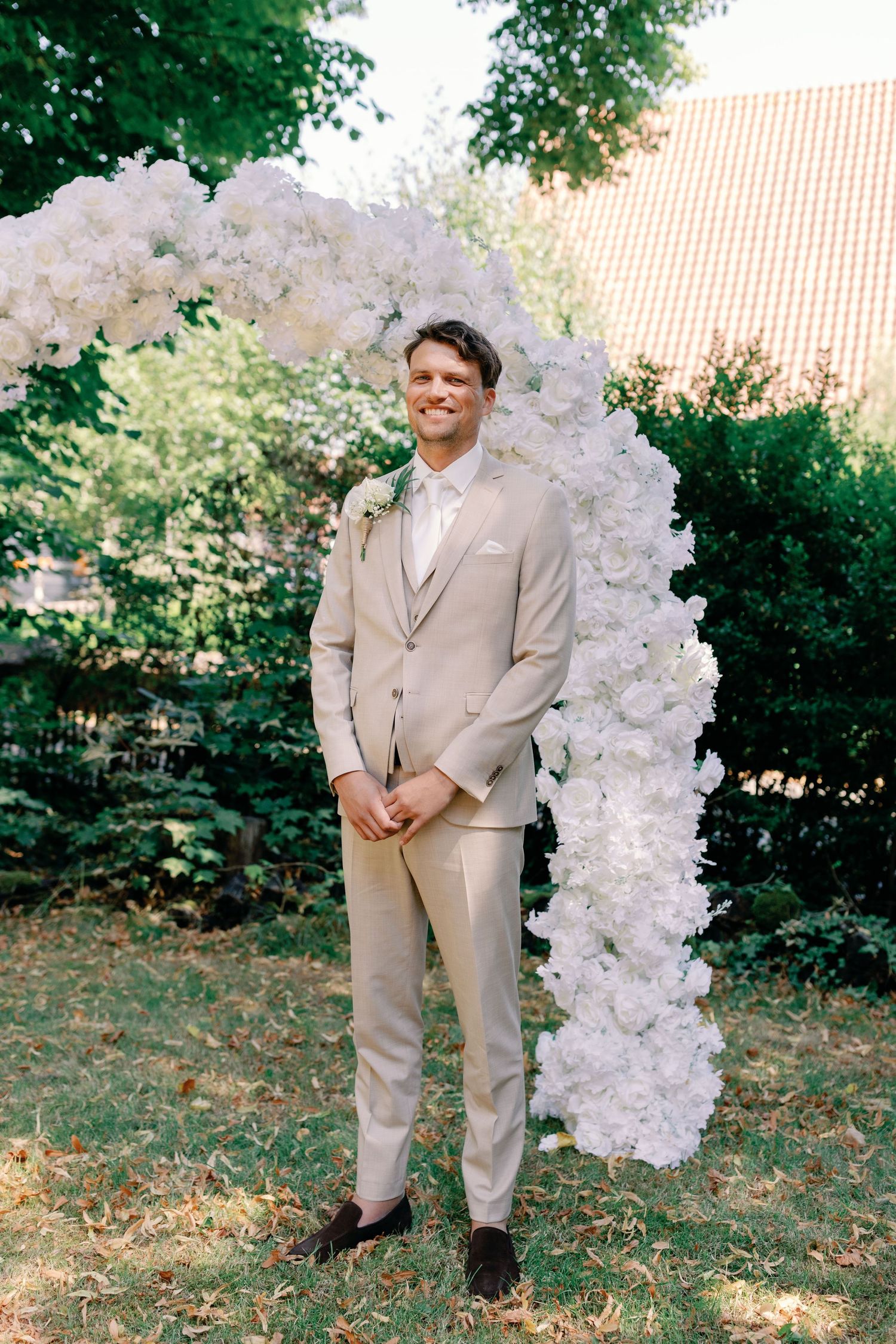 A wedding arch made of white flowers stands in a lush garden setting with a person in a beige suit nearby.