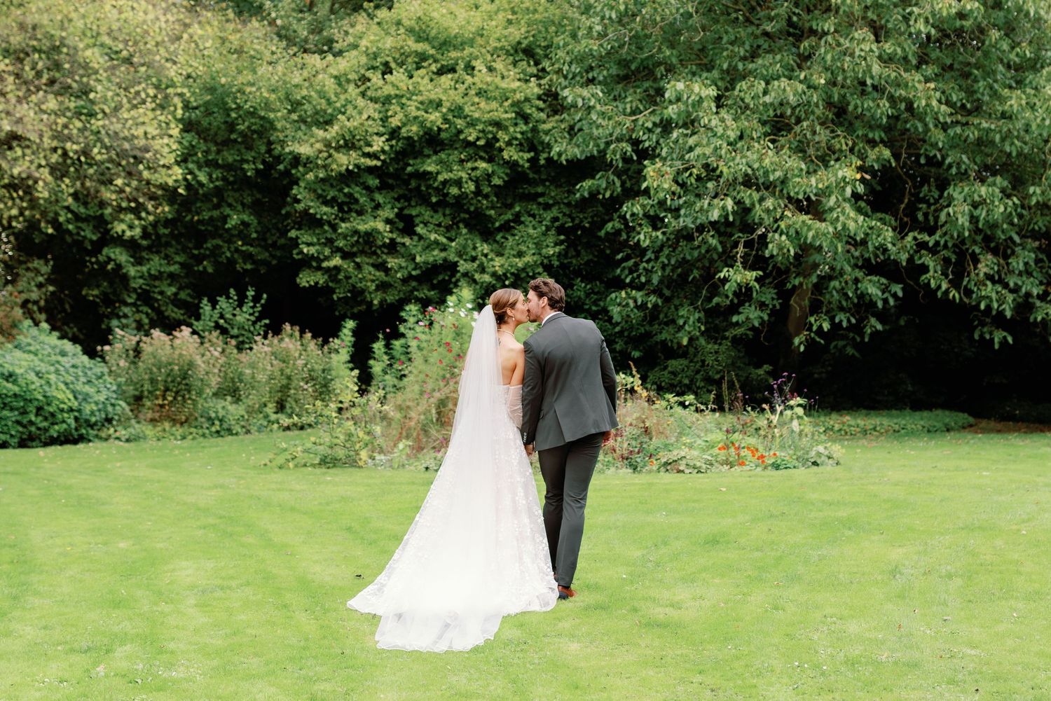 A couple in formal wedding attire embraces in a lush green garden setting.