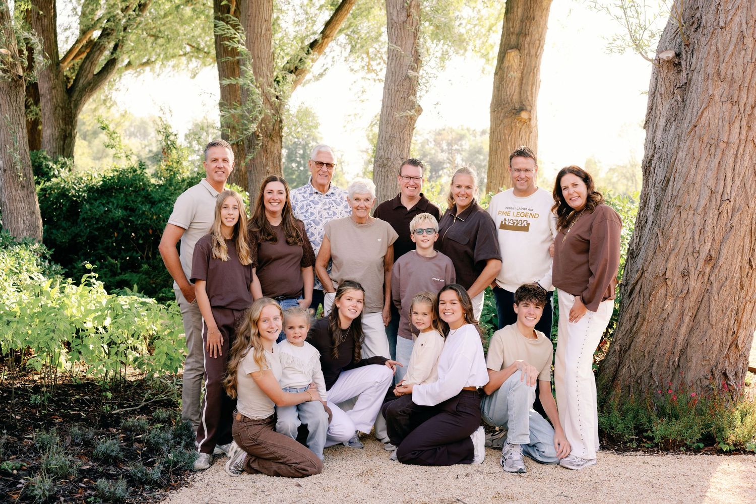 Large extended family posing together outdoors beside tall trees, with adults standing in back and children and teens seated in front.