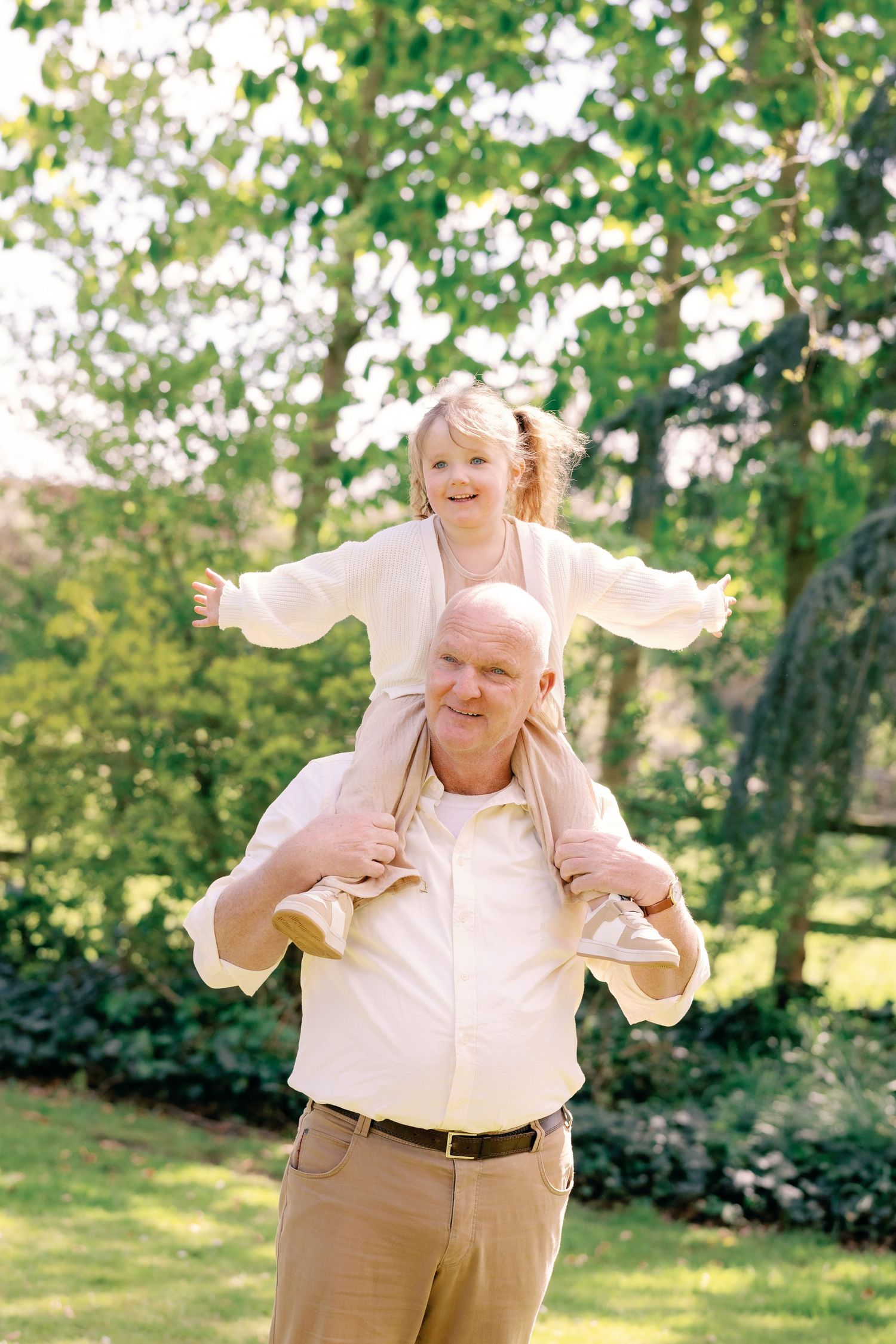 Grandfather smiling as he carries his young granddaughter on his shoulders in a sunny green park.