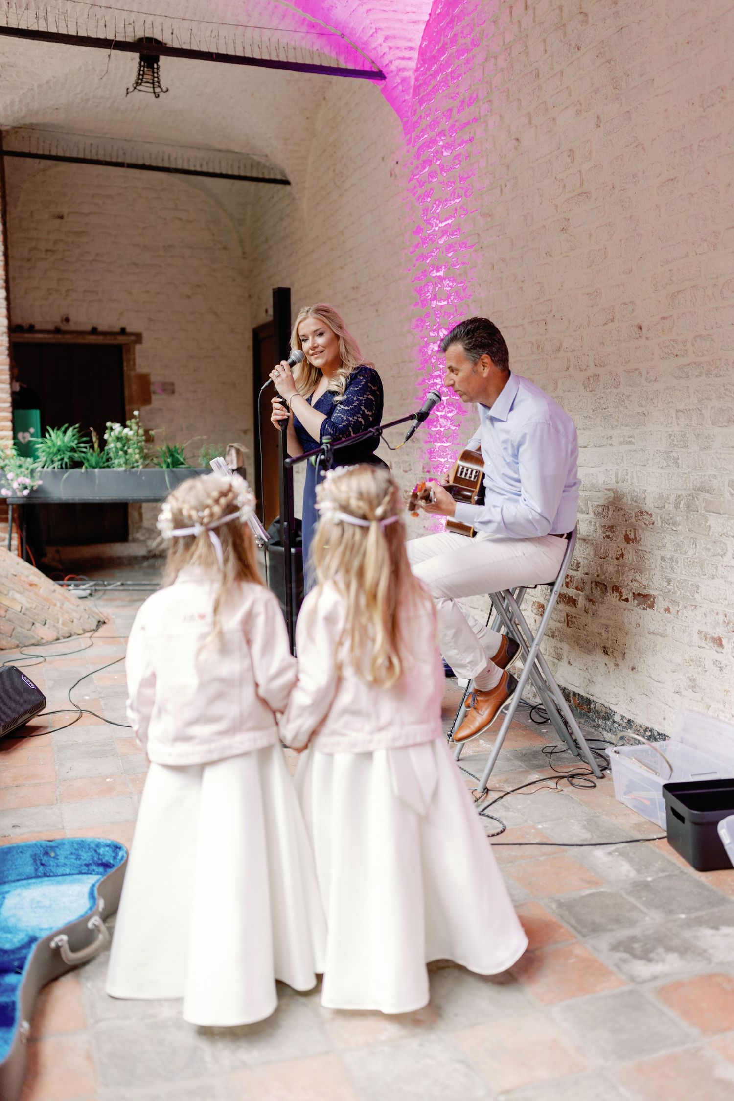 Young children in matching white dresses kneel and play together in a brick-walled space with pink lighting.