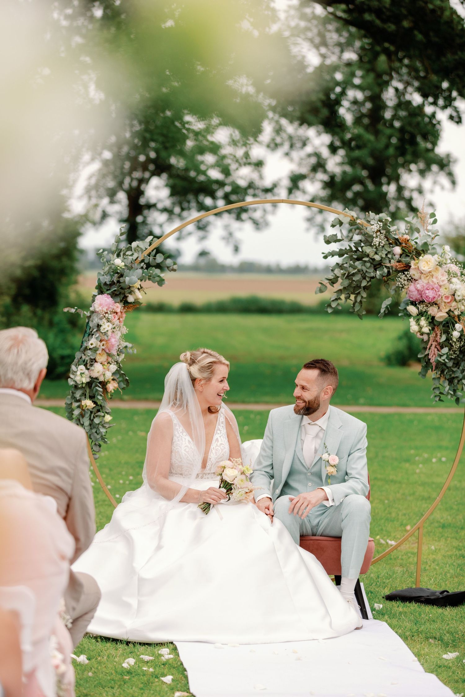 A romantic wedding couple shares a joyful moment while sitting on a blanket under blooming rose arches in a garden setting.