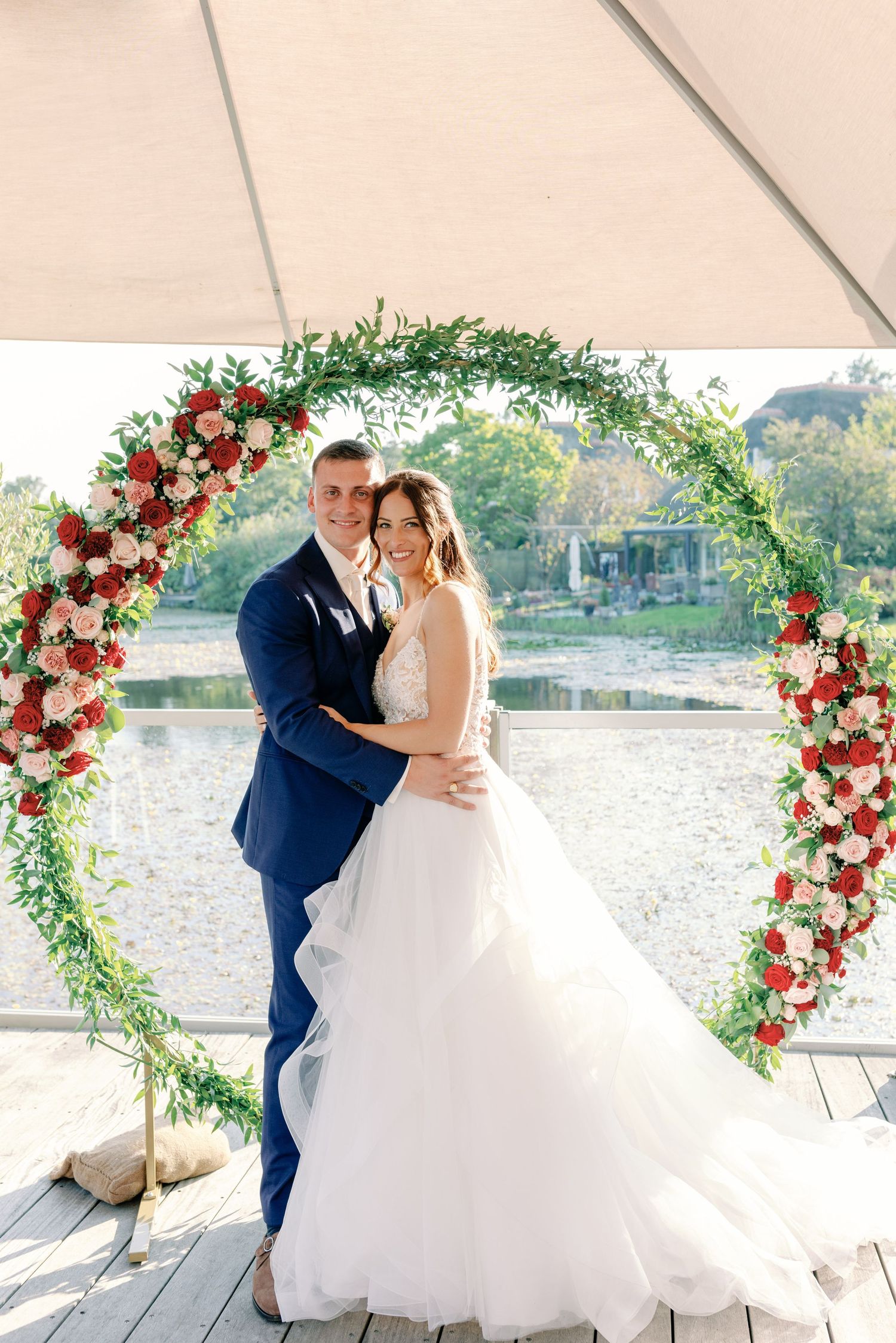 Bride in flowing white gown poses with groom under floral arch by waterfront.
