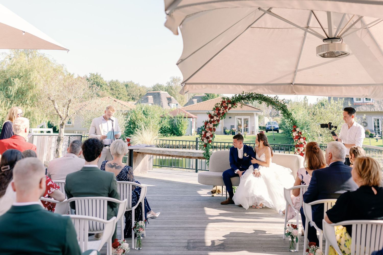A scenic outdoor wedding ceremony with guests seated under a canopy and floral arch.