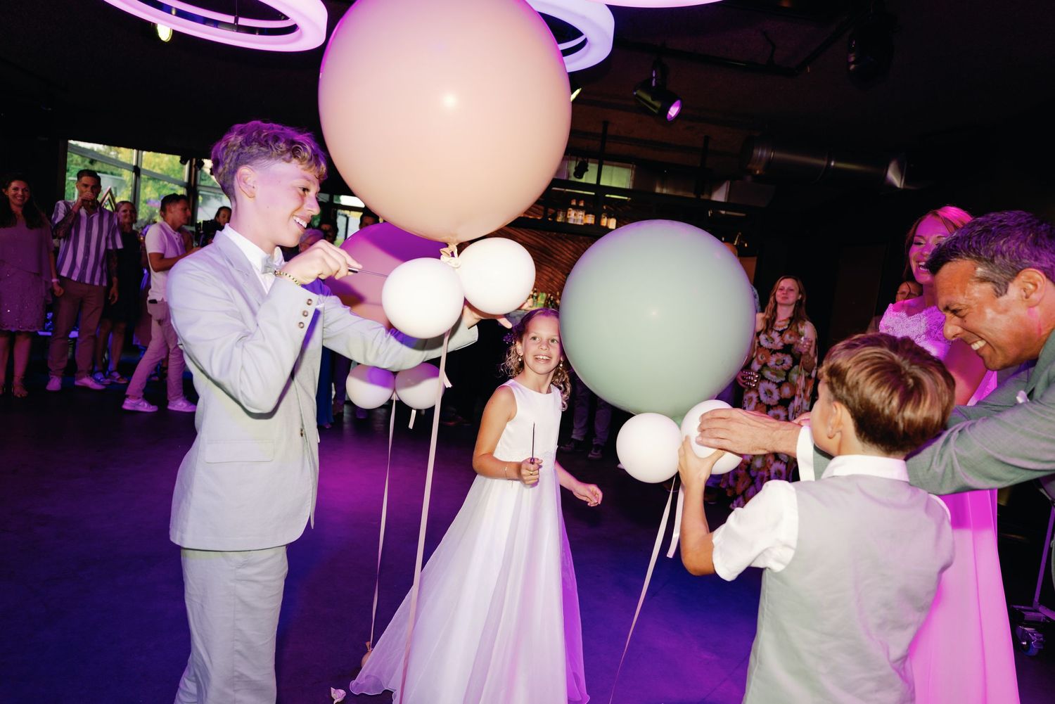 Wedding guests dance and celebrate with giant white balloons under purple lighting at an evening reception.