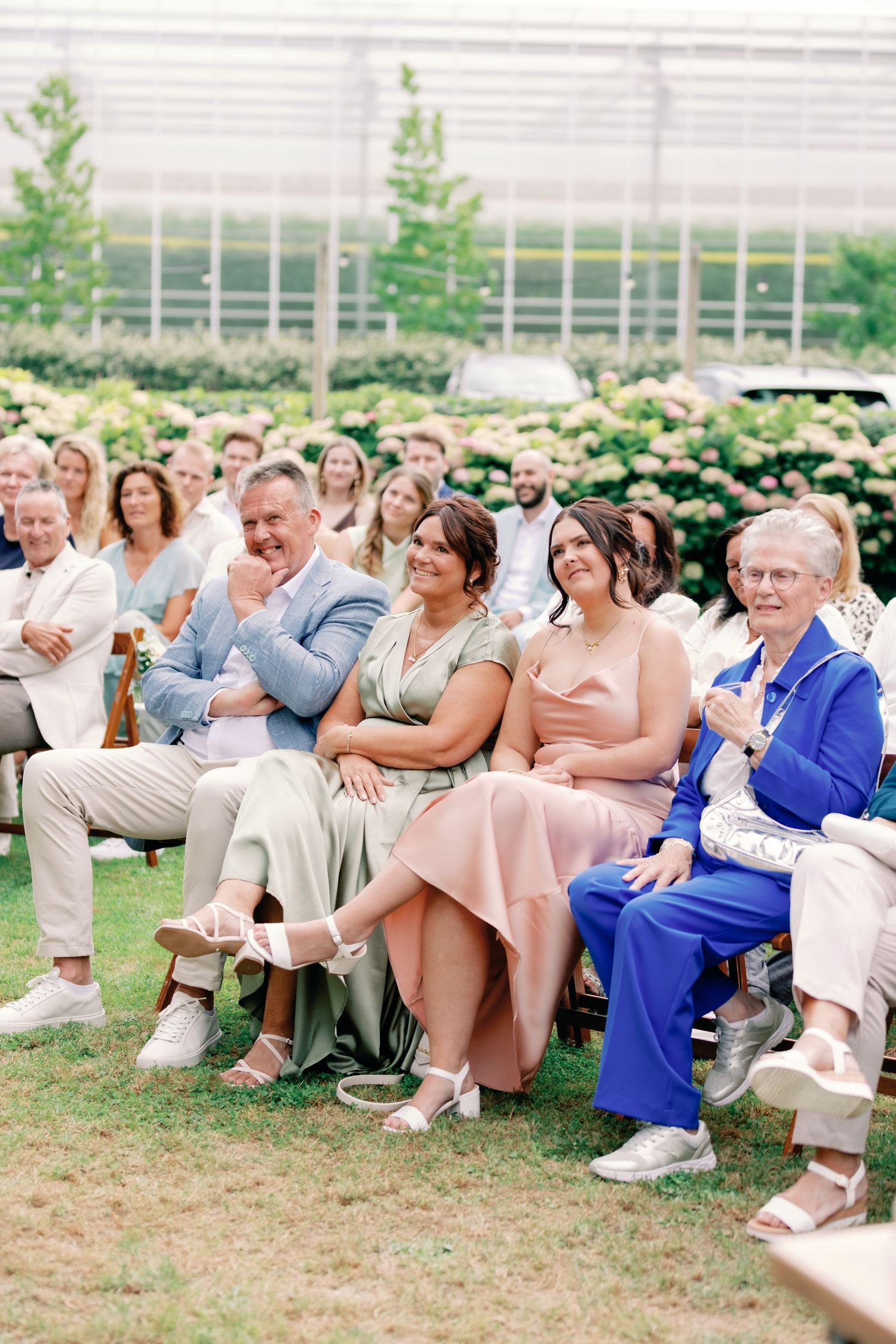 Wedding guests seated on white chairs outdoors watch ceremony in a garden setting with greenhouse in background.
