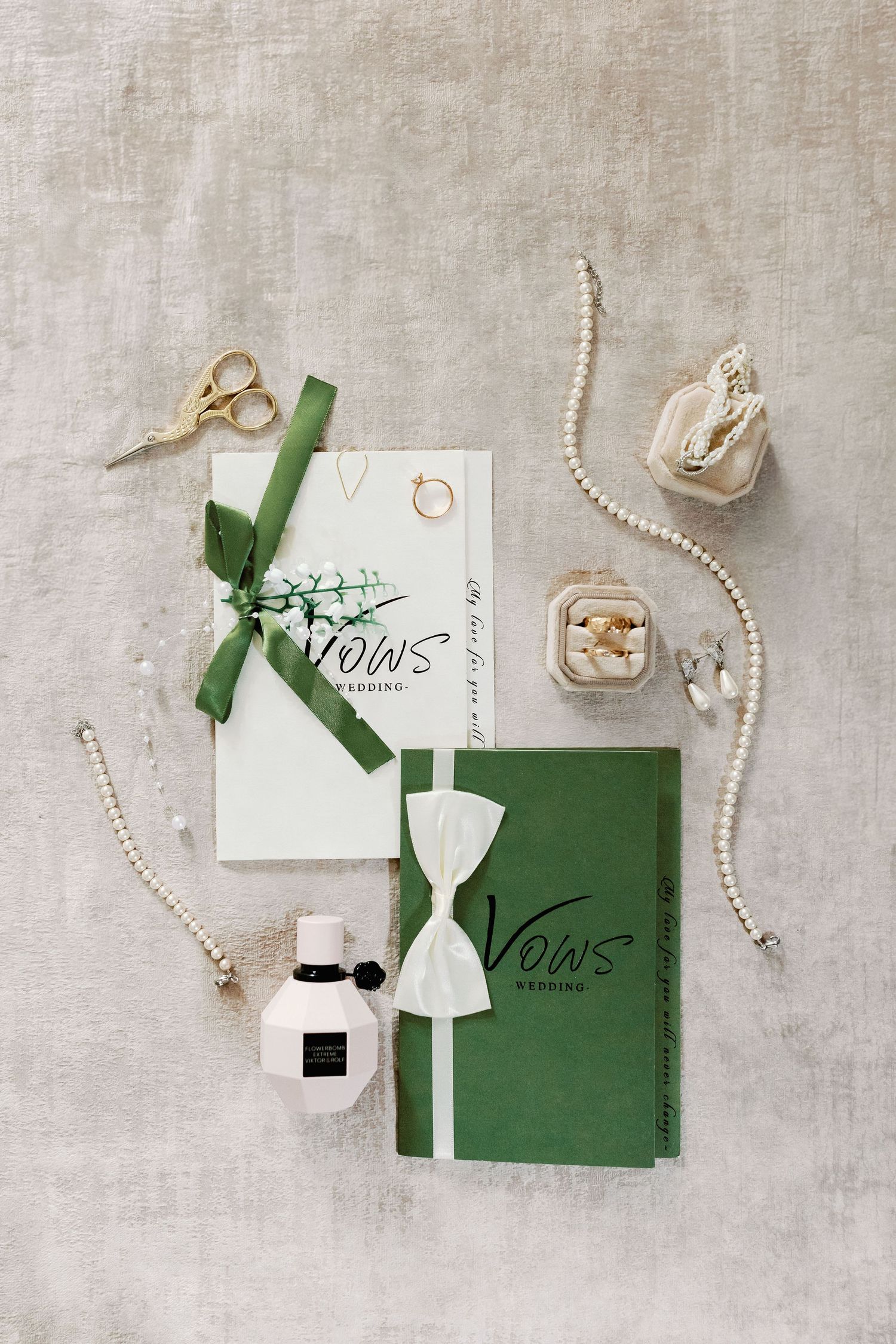 Elegant wedding invitation flatlay with green envelopes, pearl necklace, and gold rings on light textured background.