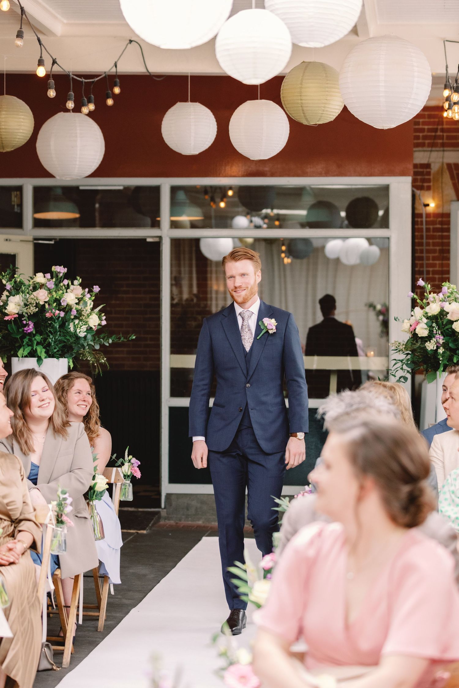 Wedding party laughs together under white paper lanterns at an indoor venue decorated with floral arrangements.