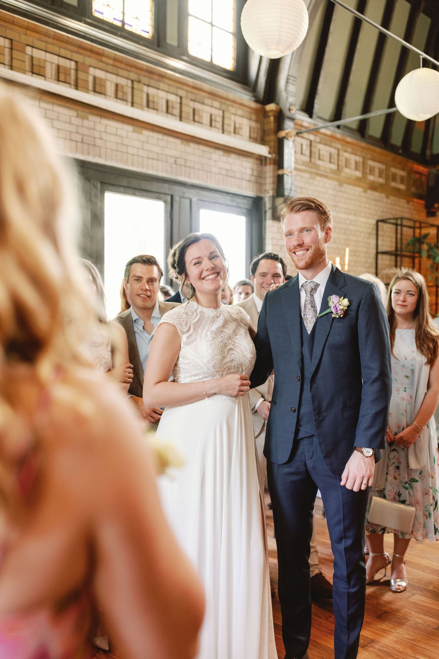 Wedding guests stand in a rustic brick venue with paper lanterns hanging from wooden beams during a celebration.