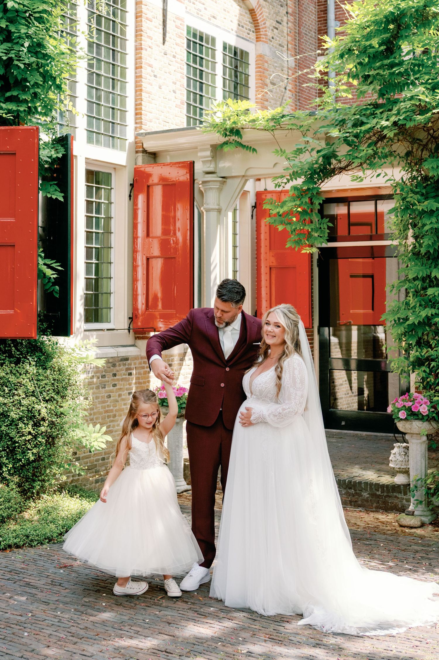 A wedding party in formal attire poses together outside a historic brick building with red shutters and green ivy.