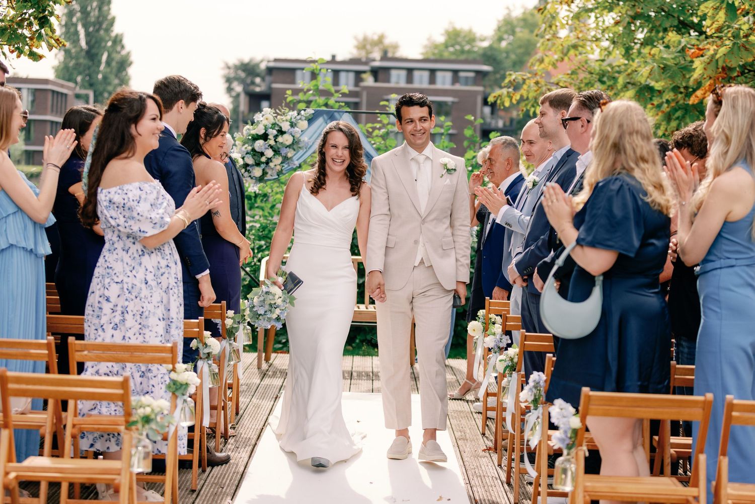 Wedding ceremony recessional with guests in blue dresses lining the outdoor aisle with wooden chairs.