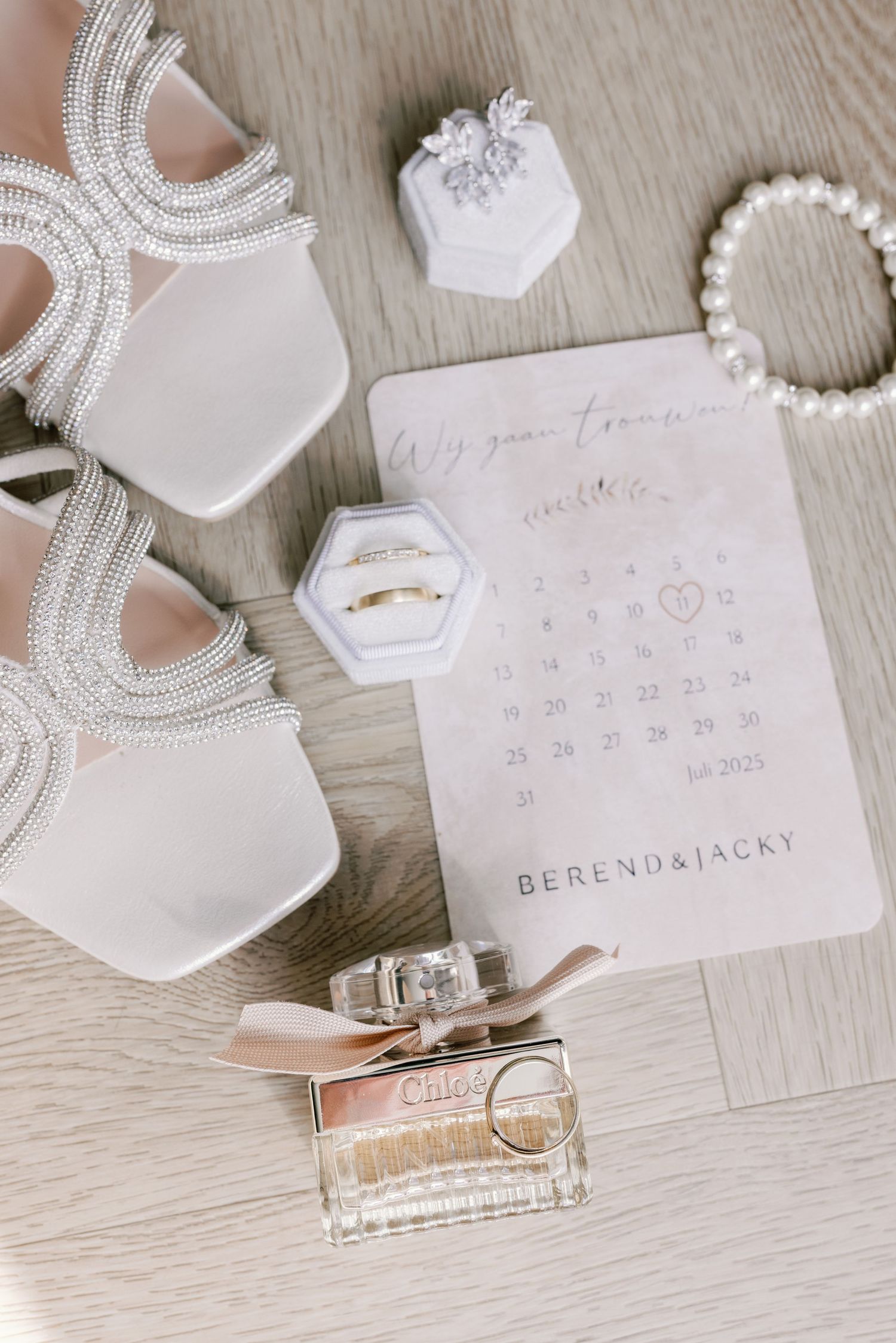 Wedding accessories including pearl necklace, calendar, perfume bottle and invitation card arranged on light wooden surface.