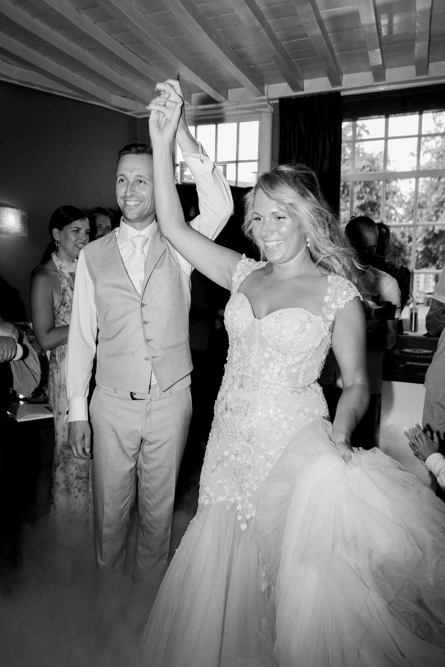 Black and white photo of a bride and groom dancing joyfully at their wedding reception, hands raised.