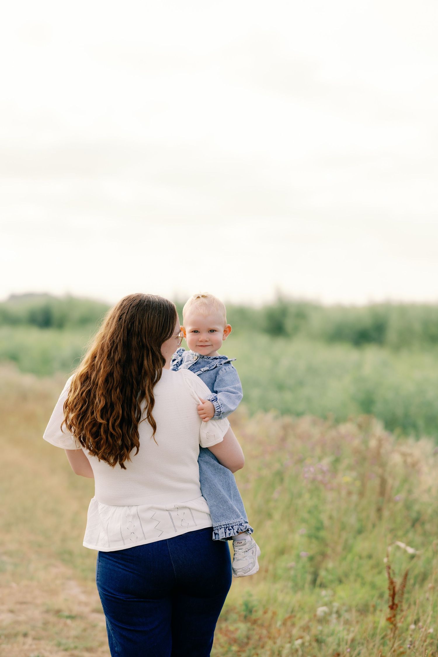 A mother holds her smiling baby in a green field, both facing away, wearing casual clothes on a cloudy day.