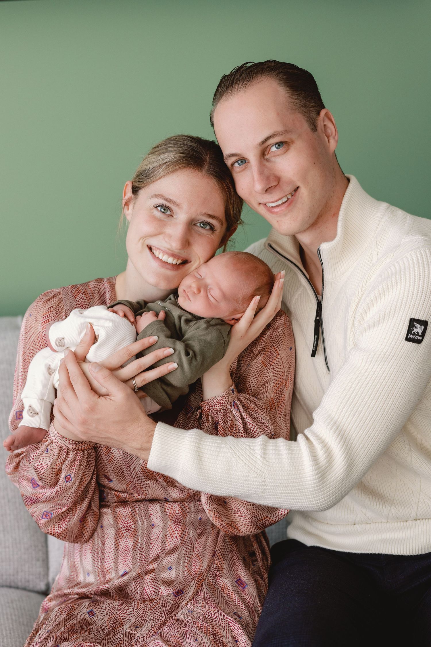 A couple in white and pink sweaters share tender moments while holding their newborn baby.