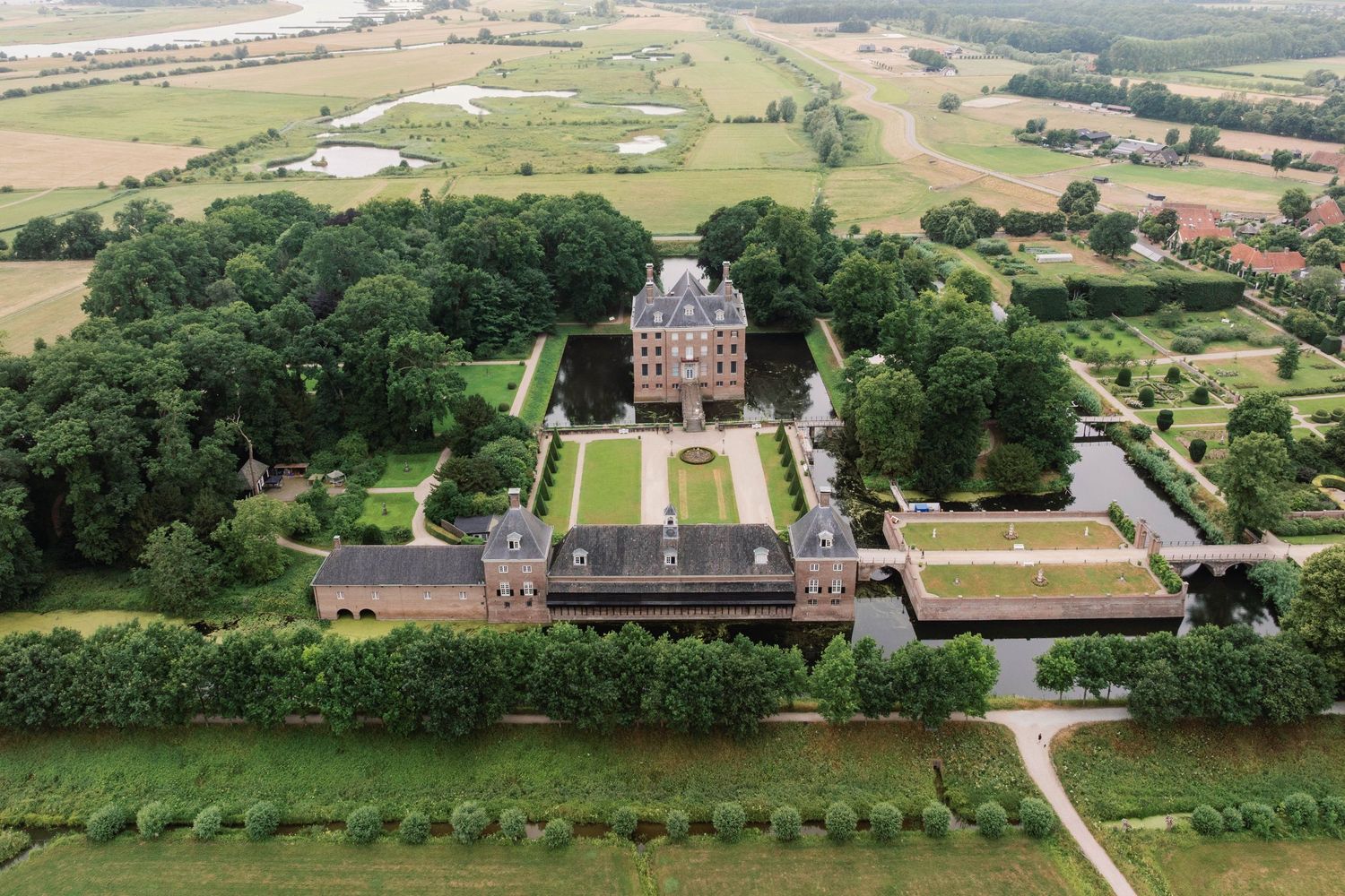 Aerial view of a grand historic mansion with manicured gardens and symmetrical layout surrounded by countryside landscape.