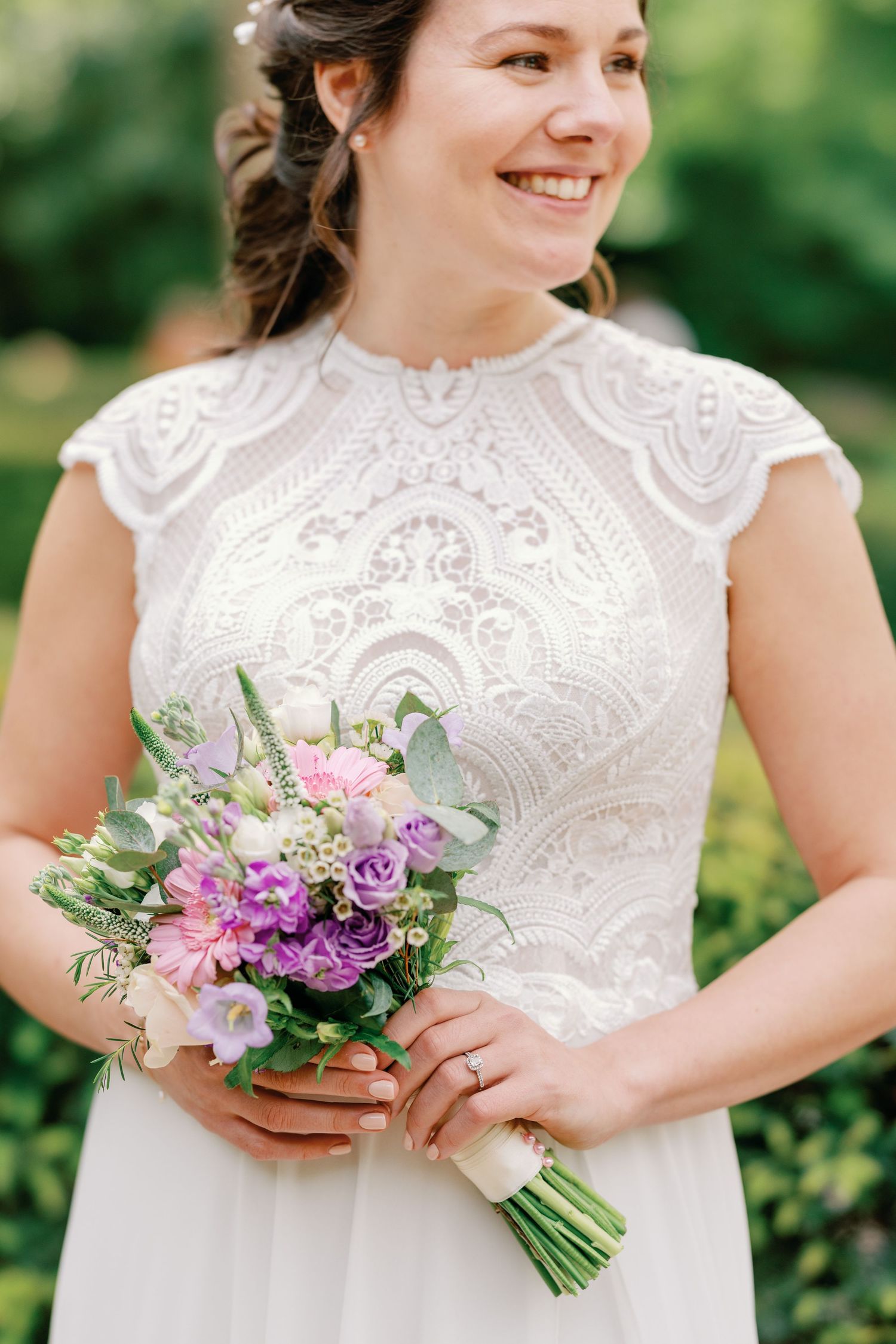 A bride in a lace cap sleeve wedding dress holds a purple and pink bouquet against a blurred green background.