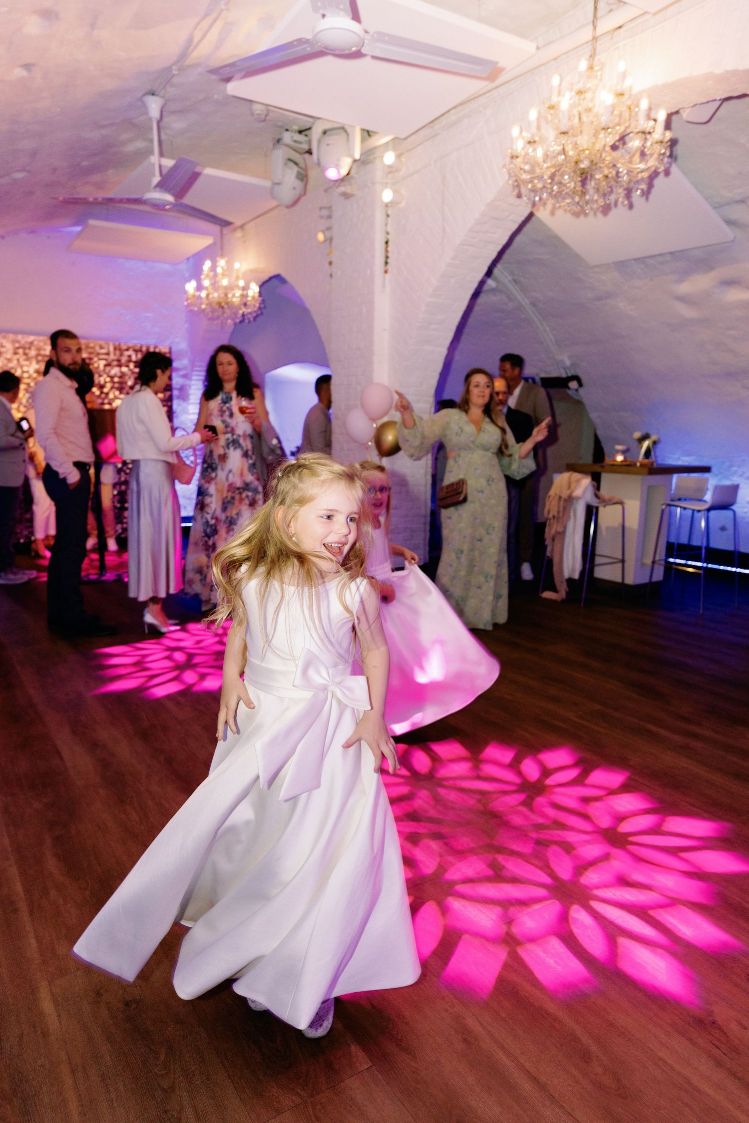 A child in a white dress twirls on a dance floor illuminated by bright pink lighting patterns and crystal chandeliers.