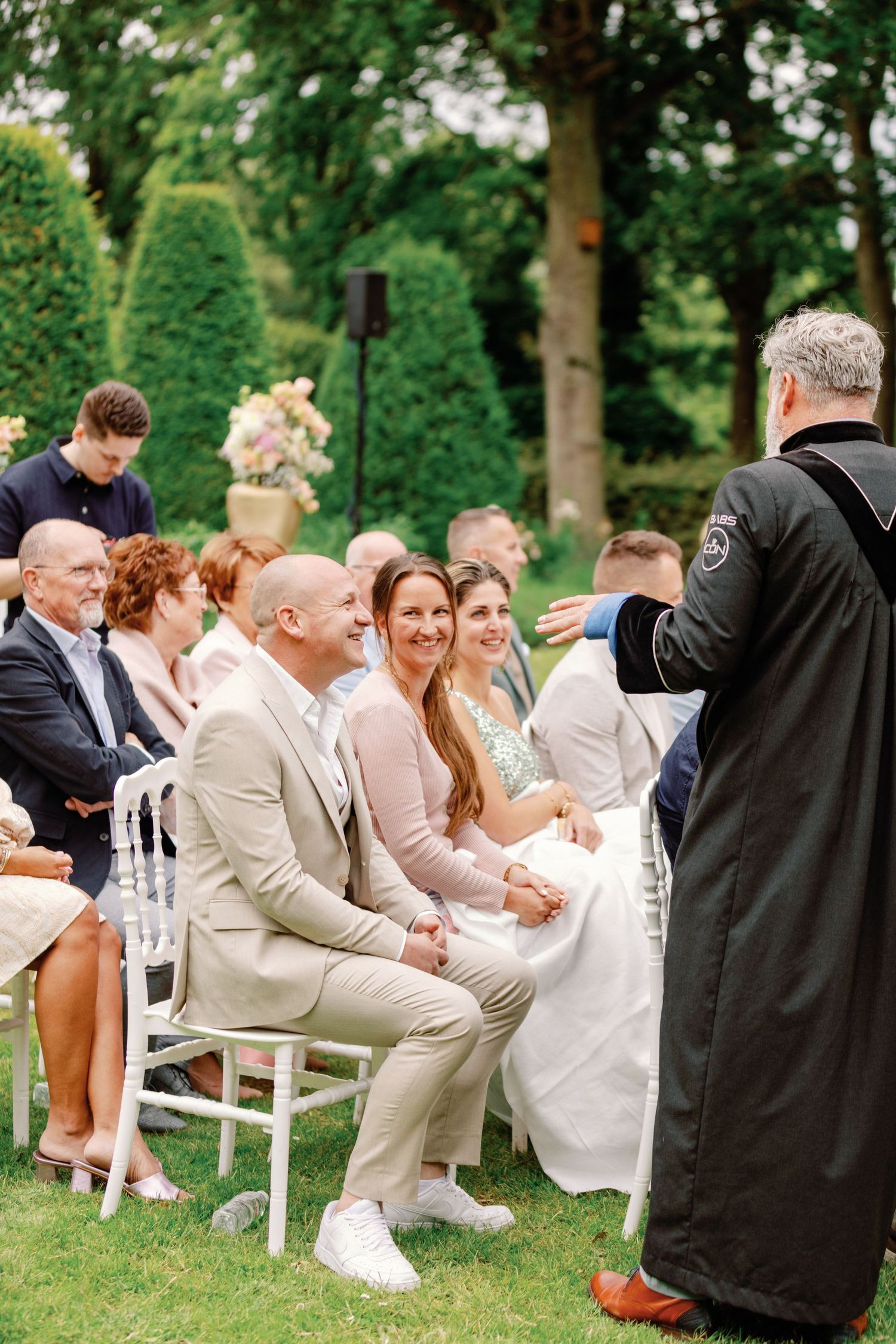 Wedding guests seated on white chairs on a lawn while an officiant conducts an outdoor ceremony.
