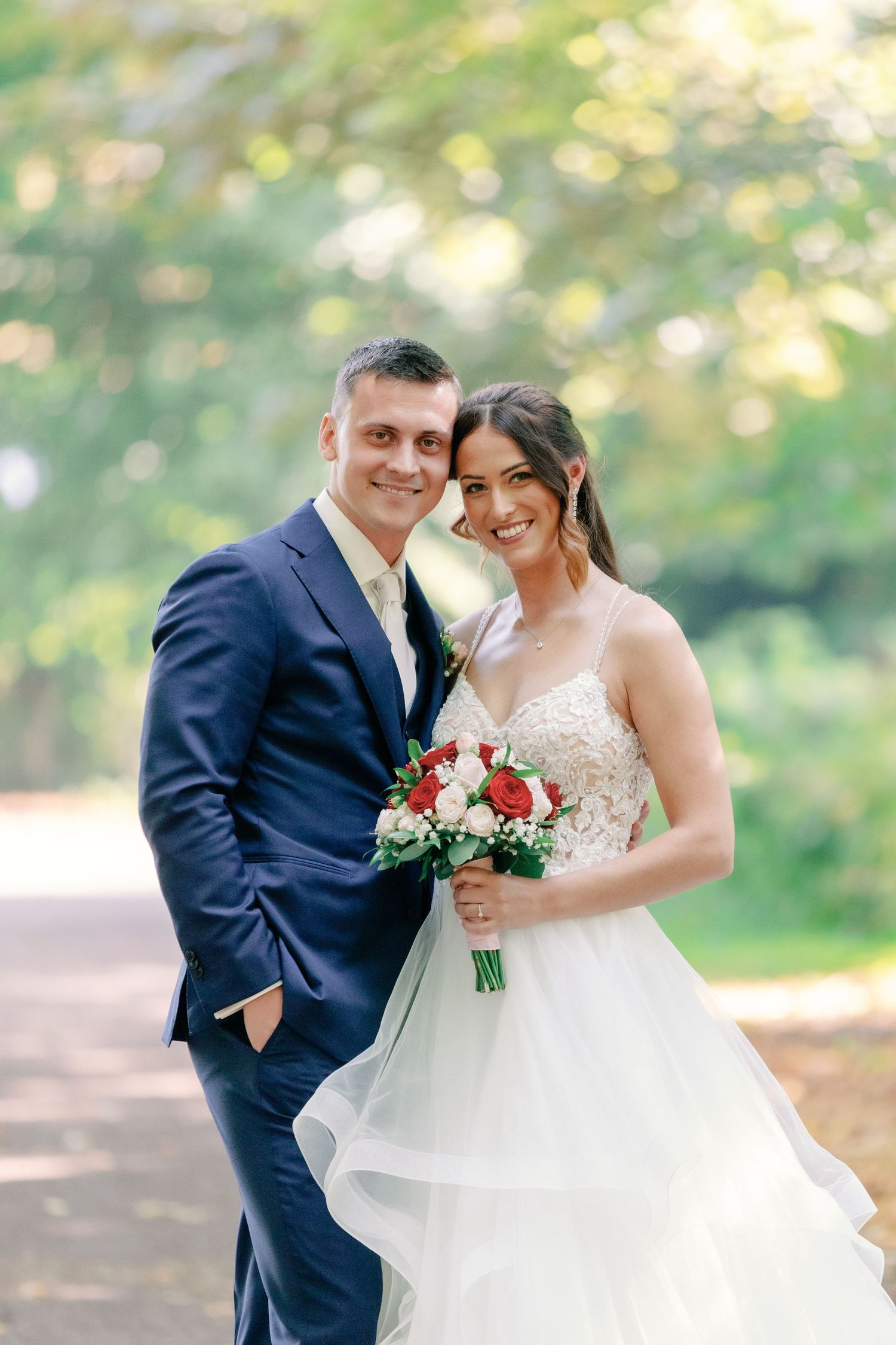 Romantic wedding portrait of couple with red bouquet in sunlit wooded setting.