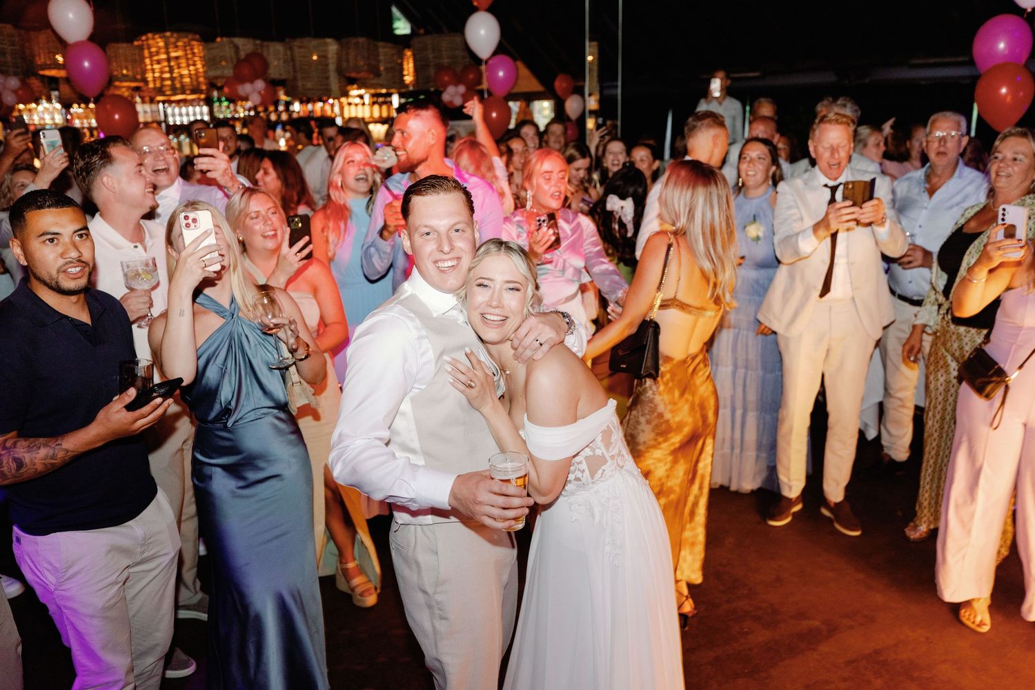 Wedding guests dance and celebrate on a dimly lit dance floor with colorful lighting and festive atmosphere.