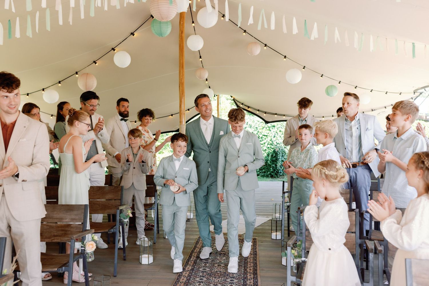 A wedding celebration under a tent with string lights and white decor as guests in light colored attire applaud.
