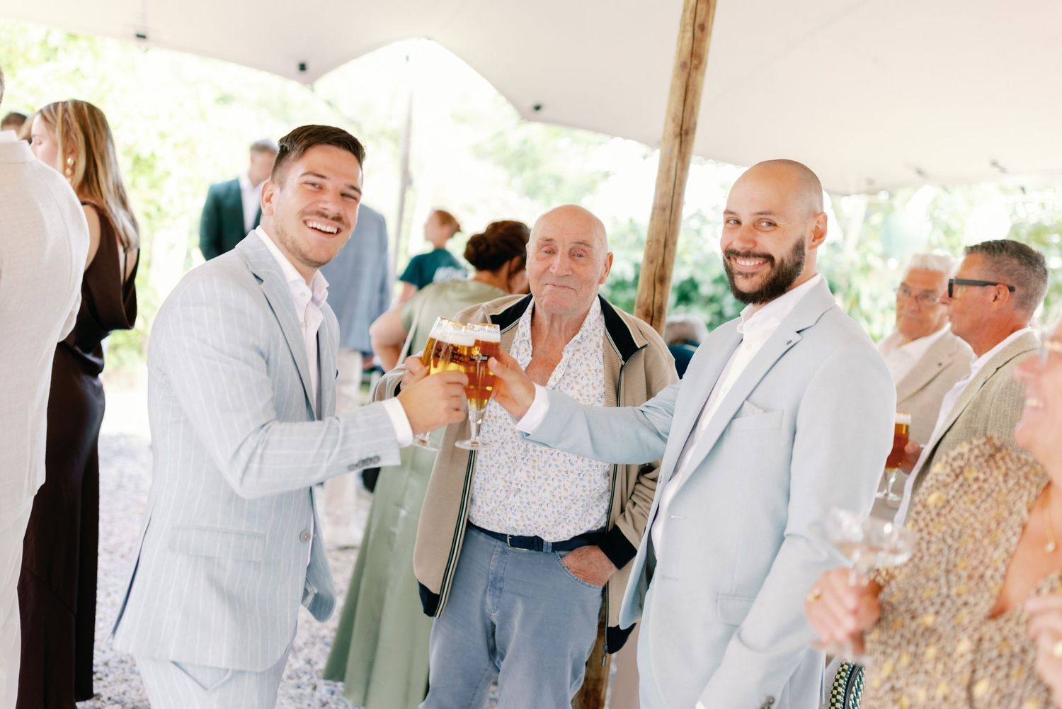 Wedding guests in light summer attire mingle and socialize under a white tent during an outdoor reception.