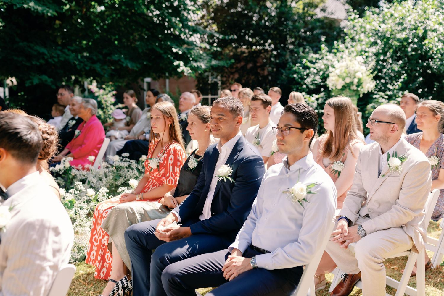 Wedding guests sit in white chairs during an outdoor garden ceremony on a sunny summer day.