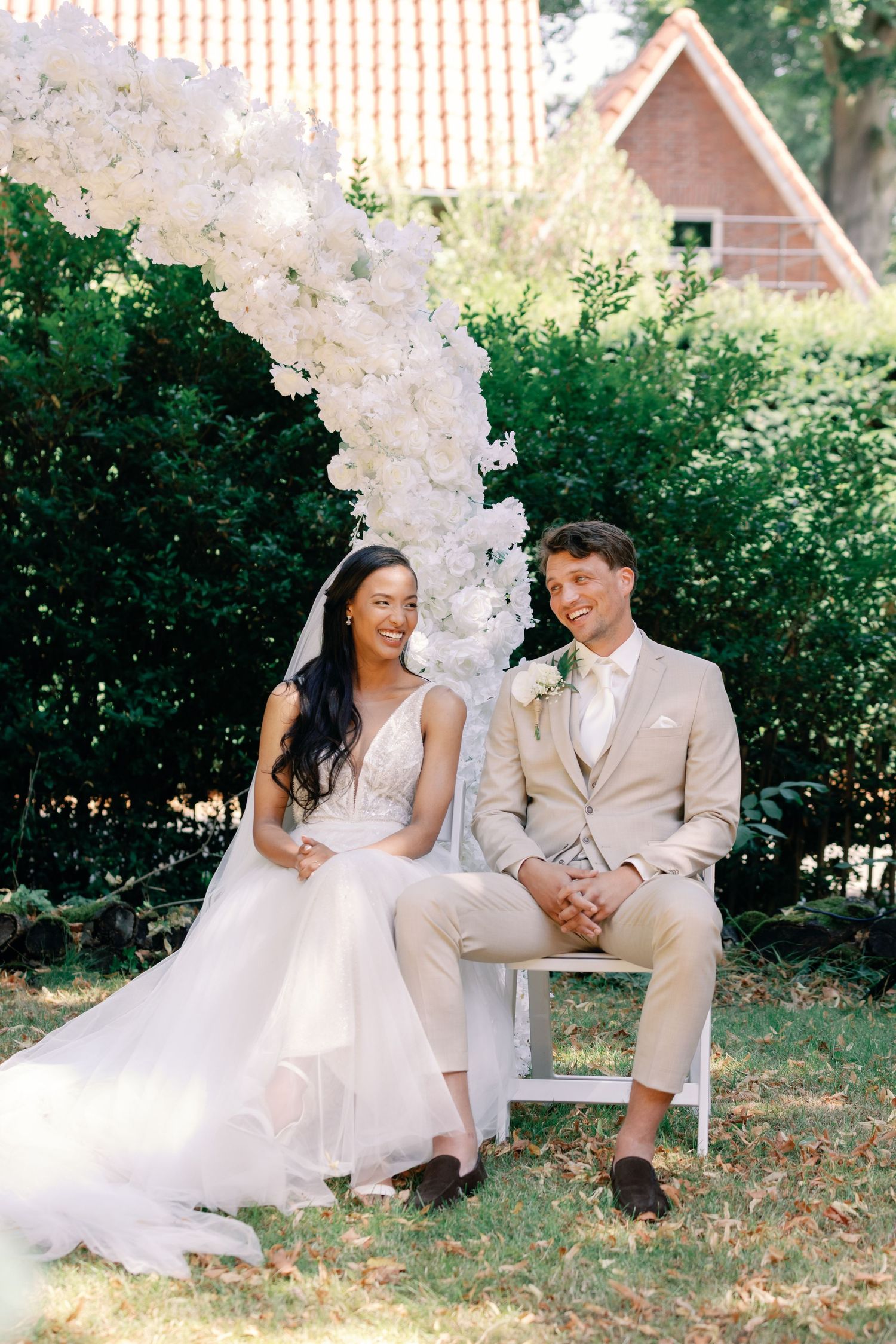 A couple shares joyful moments on a garden bench surrounded by elegant white floral arrangements and greenery.