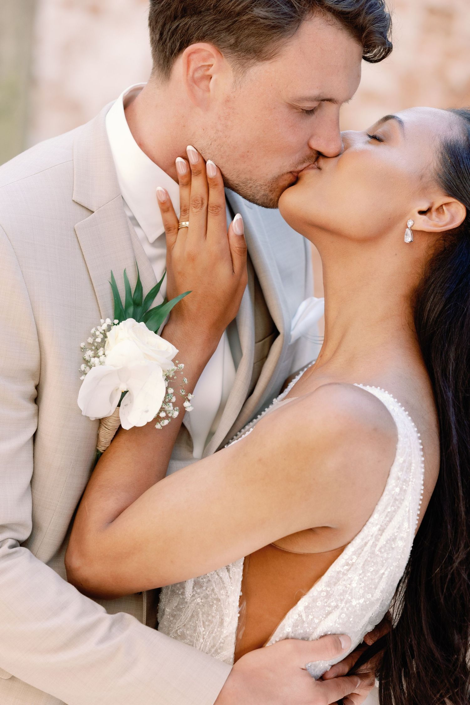 A romantic wedding couple shares a tender kiss, with the bride wearing a white gown and groom in a beige suit.