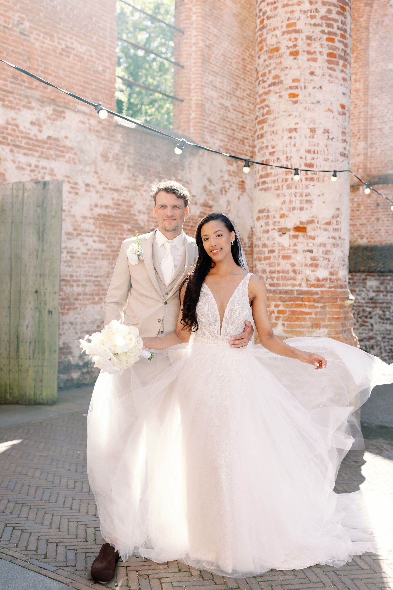 A bride in a flowing white gown twirls and dances joyfully against a rustic brick wall backdrop.