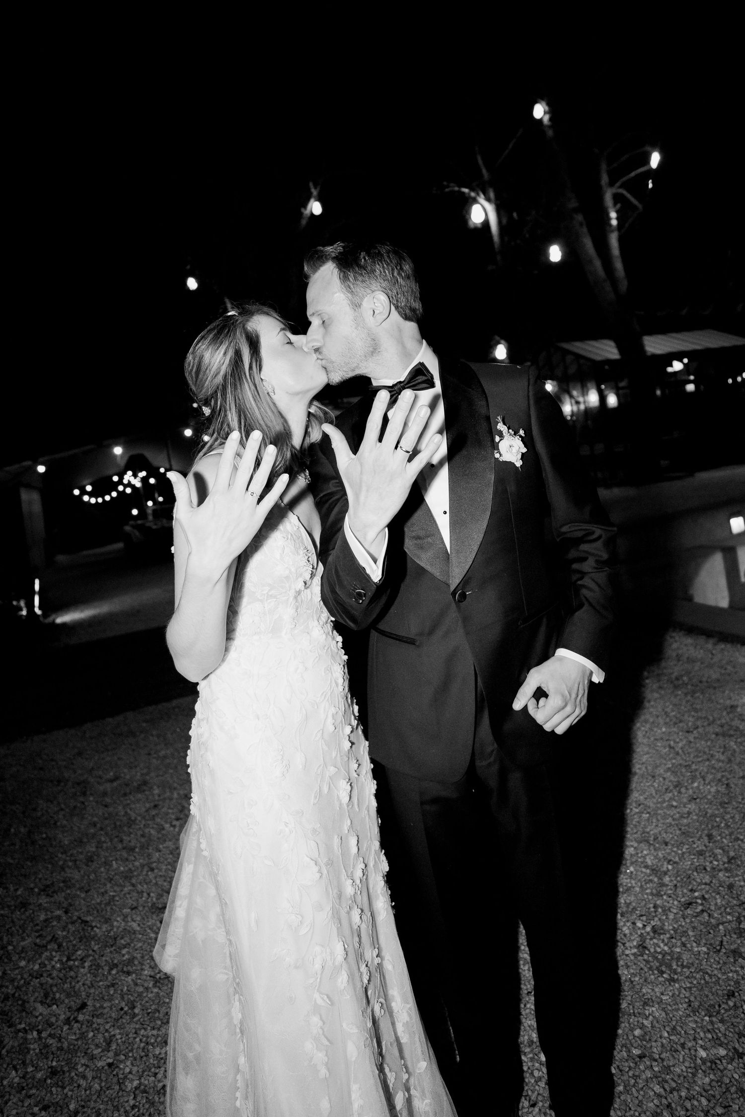 Black and white photo of a bride and groom kissing at night while showing off their wedding rings, with string lights glowing behind them.
