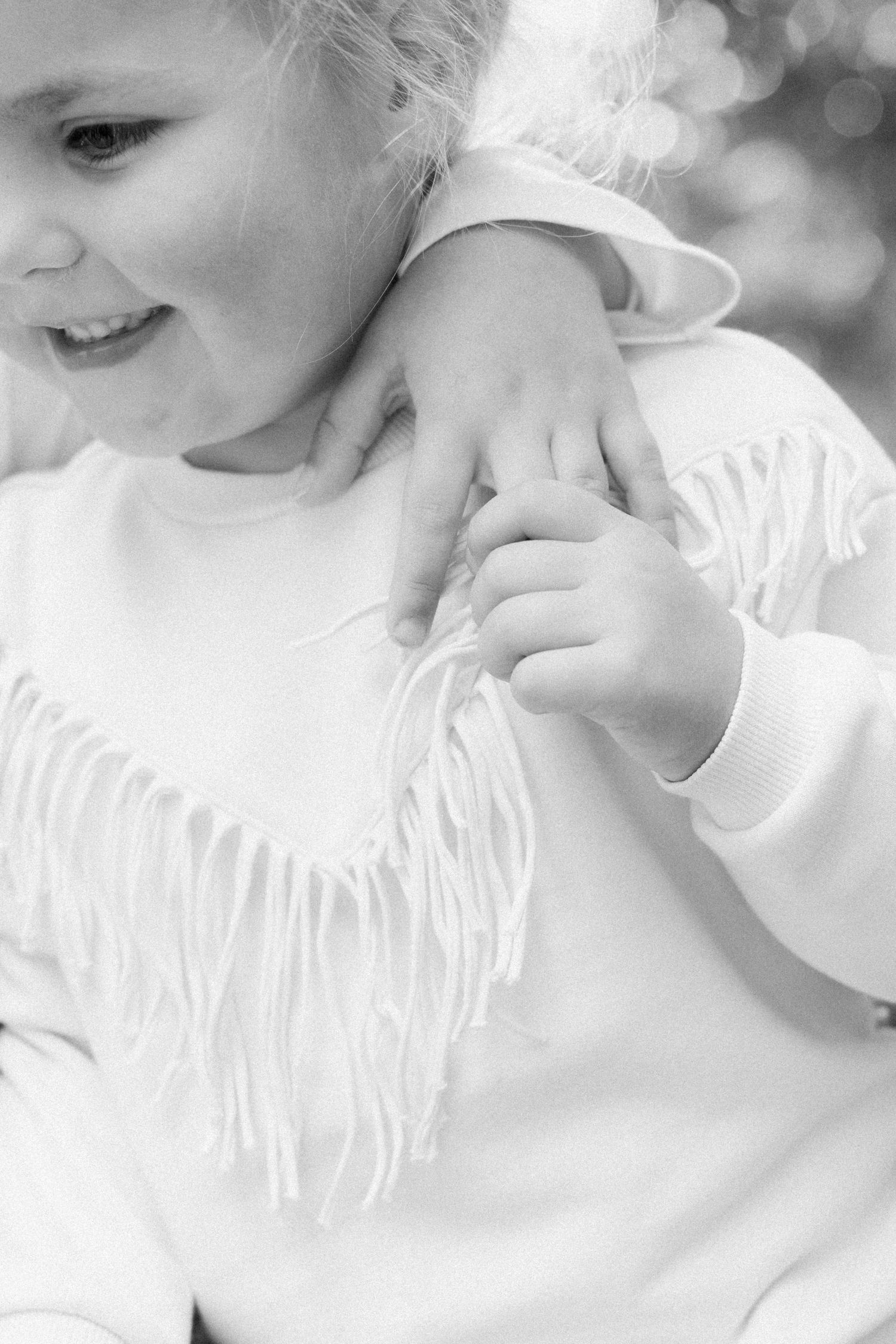 Black and white close-up of a smiling toddler in a fringed sweatshirt with a small hand resting on her shoulder.