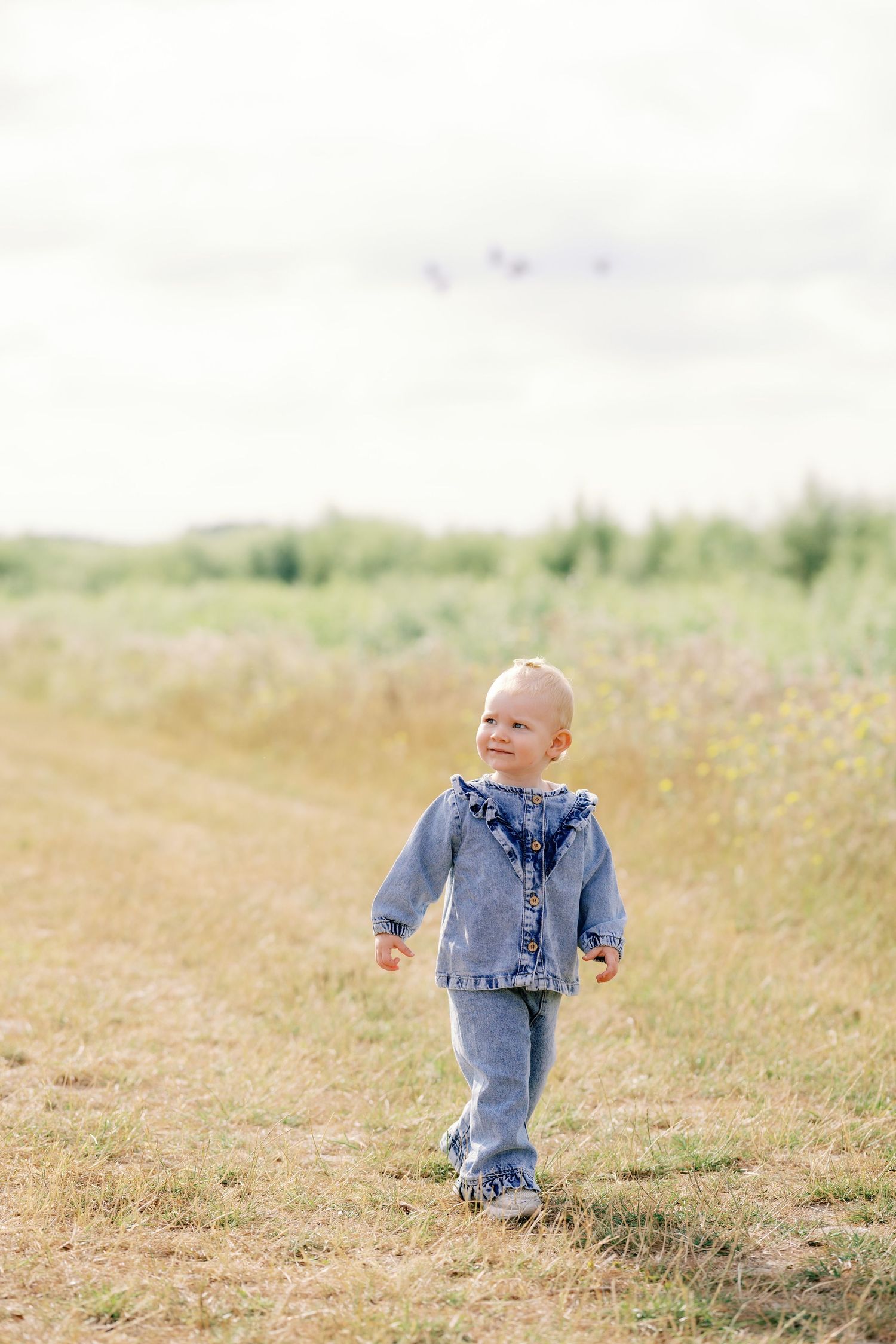 A toddler in a denim outfit walks along a rural path through a grassy field under a cloudy sky.