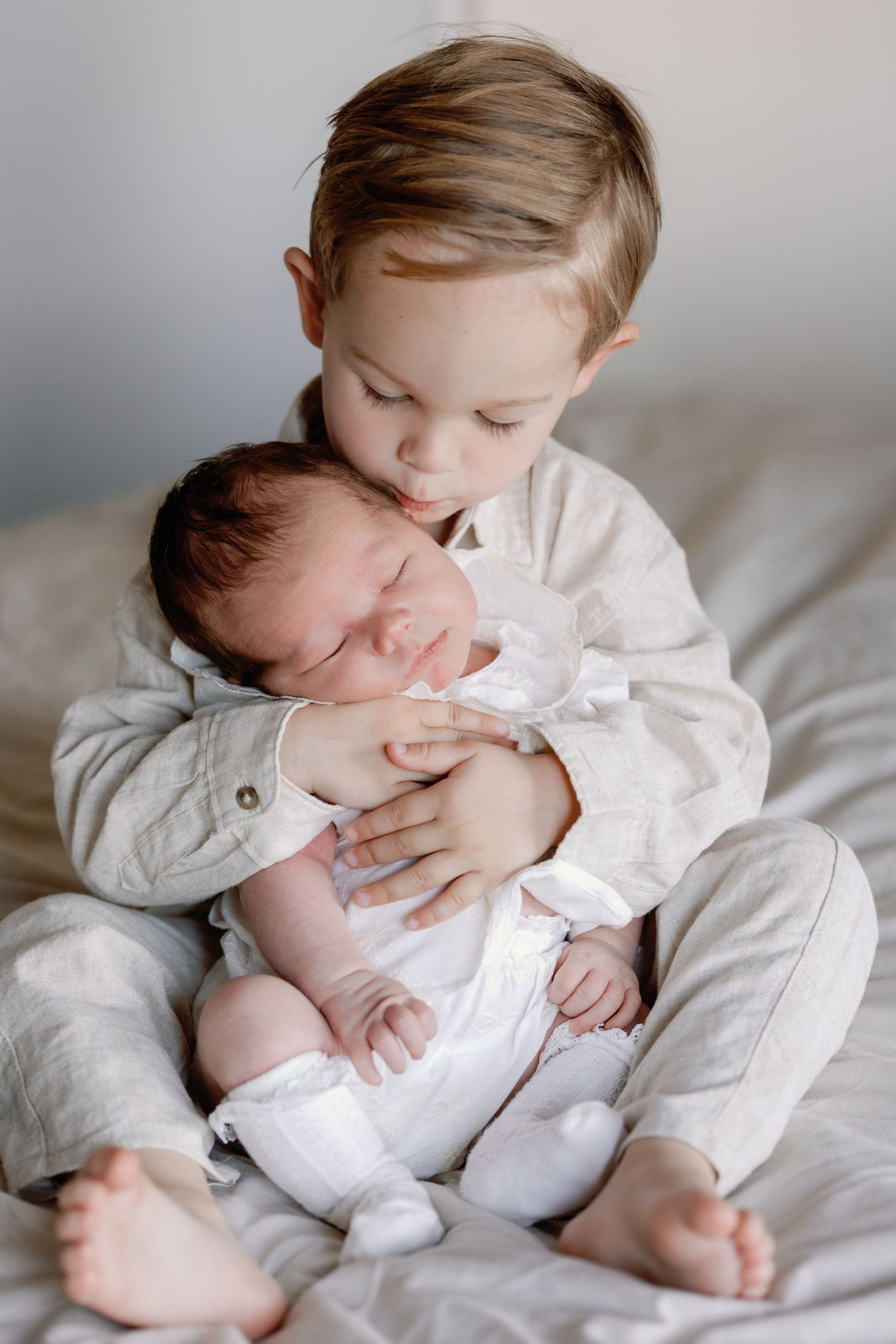 Child in grey sweater gently holding and kissing newborn sibling.