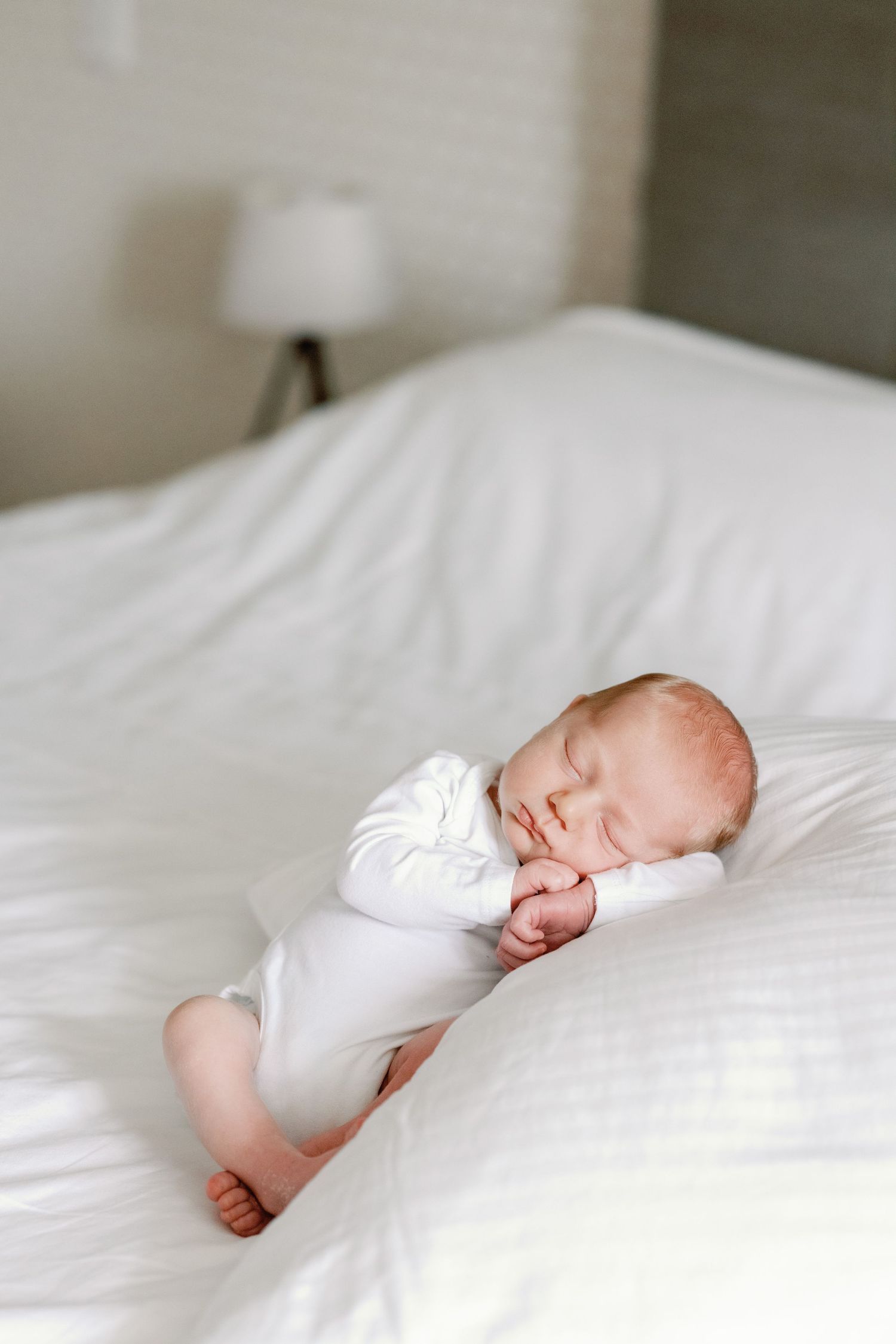 A sleeping newborn baby in white clothing rests peacefully on white bedding.
