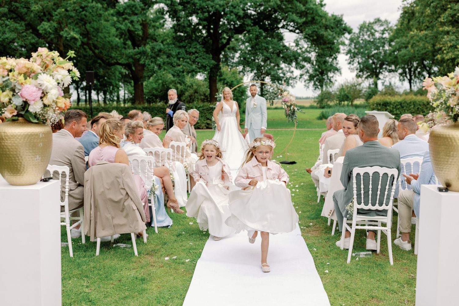 Outdoor garden party setting with white chairs and tables under lush green trees.