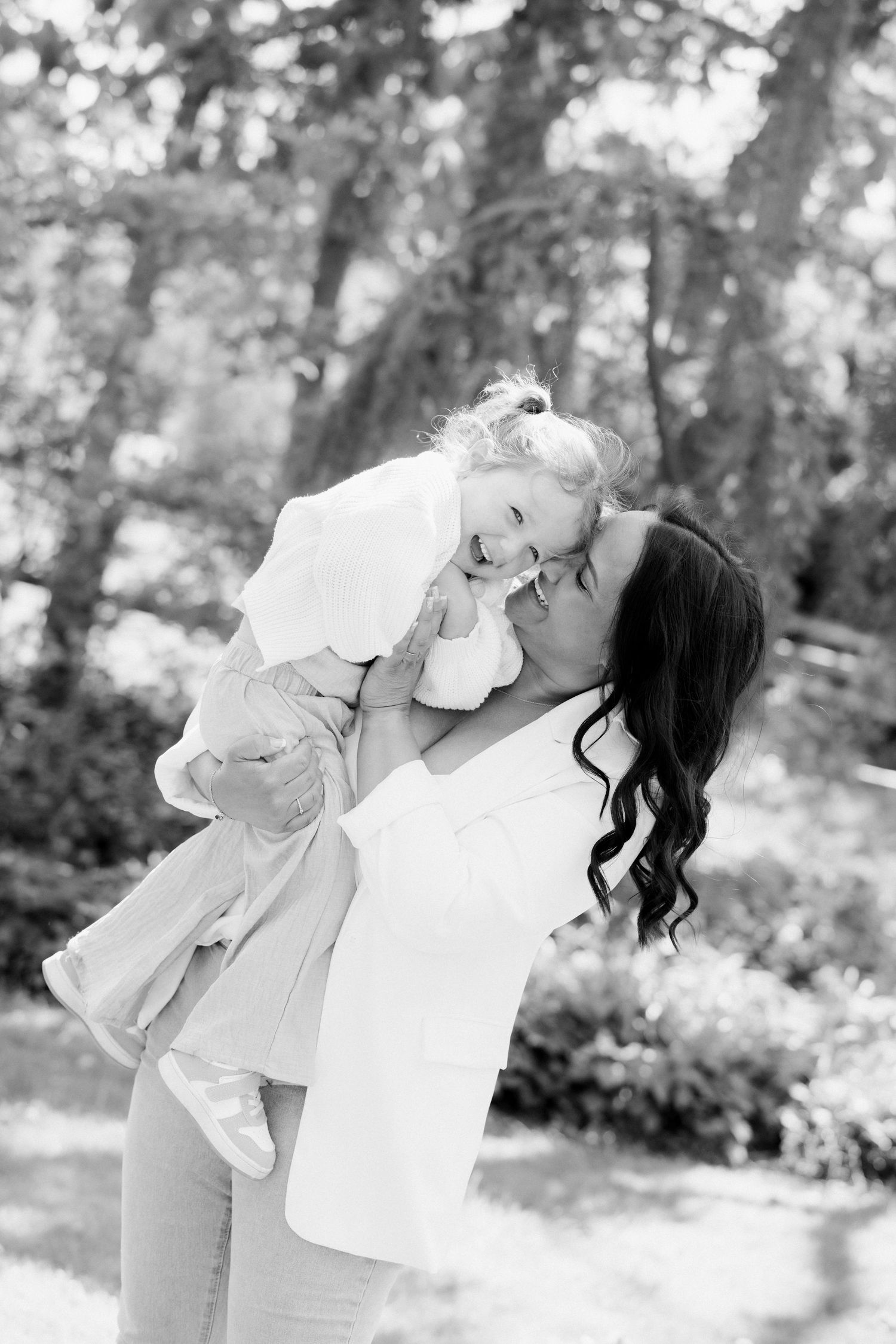 Black and white photo of a mother lifting and kissing her laughing toddler daughter outdoors in a lush green park.