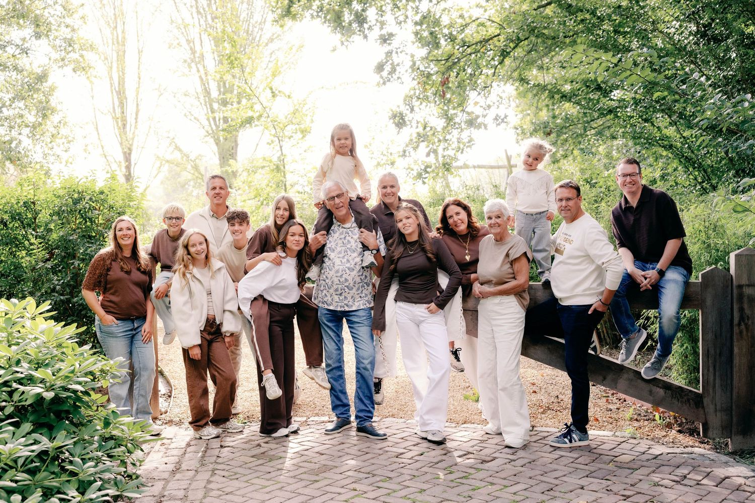 Large multigenerational family posing together on a brick path in a sunny park with green trees, children, and adults in casual outfits.