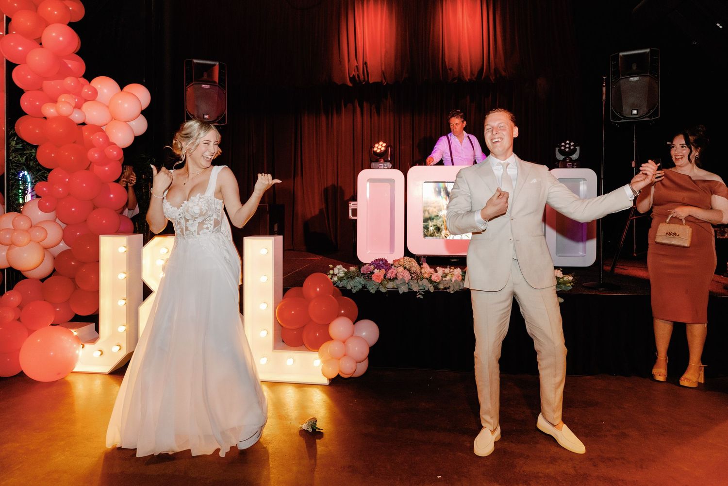 Wedding party dancing and celebrating in a ballroom decorated with pink balloons and ambient red lighting.