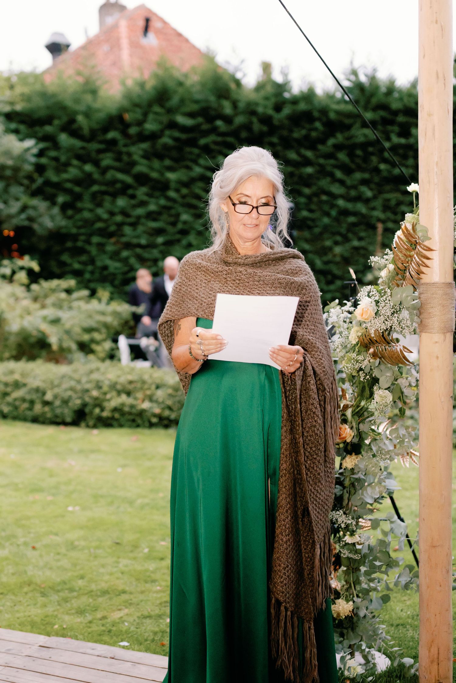 Someone wearing glasses and a brown shawl reads from a paper while standing in a garden wearing an emerald green skirt.