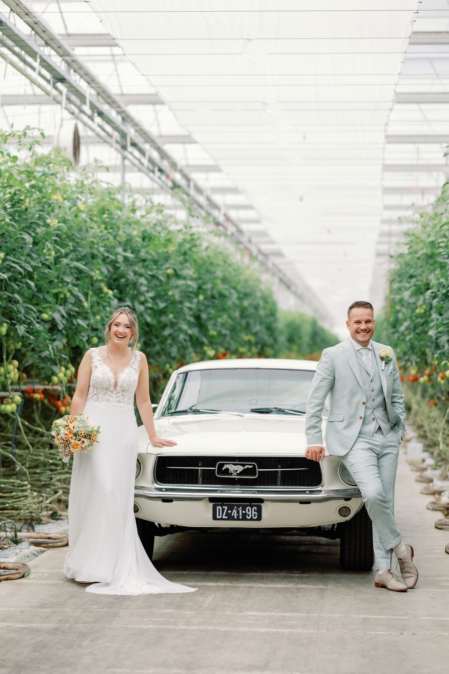 A couple poses with a classic Ford Mustang inside a greenhouse with lush green plants lining both sides.